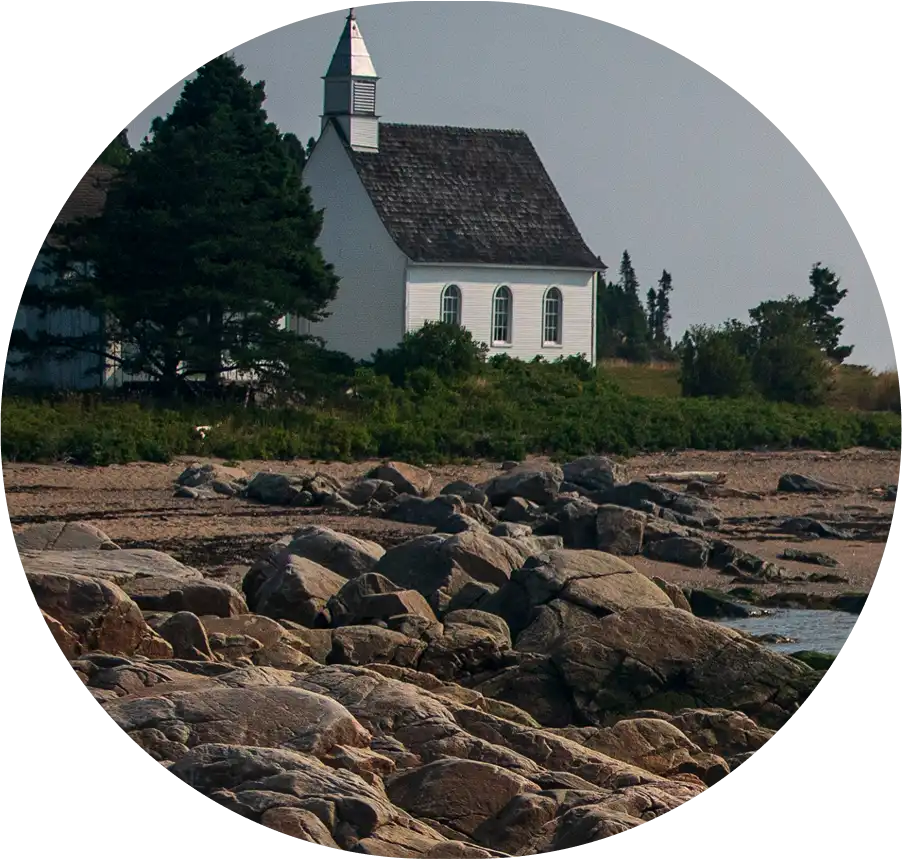 Petite église blanche en bord de mer, entourée de conifères, vue depuis un rivage rocheux sous une lumière douce.