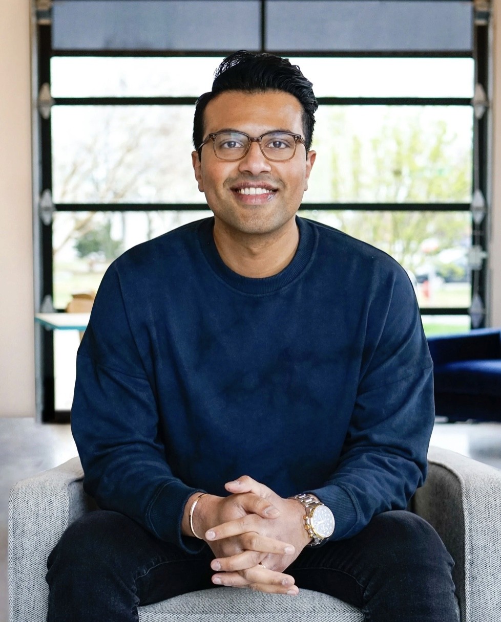 Smiling young man with curly hair, wearing a dark hoodie, against a softly lit background.