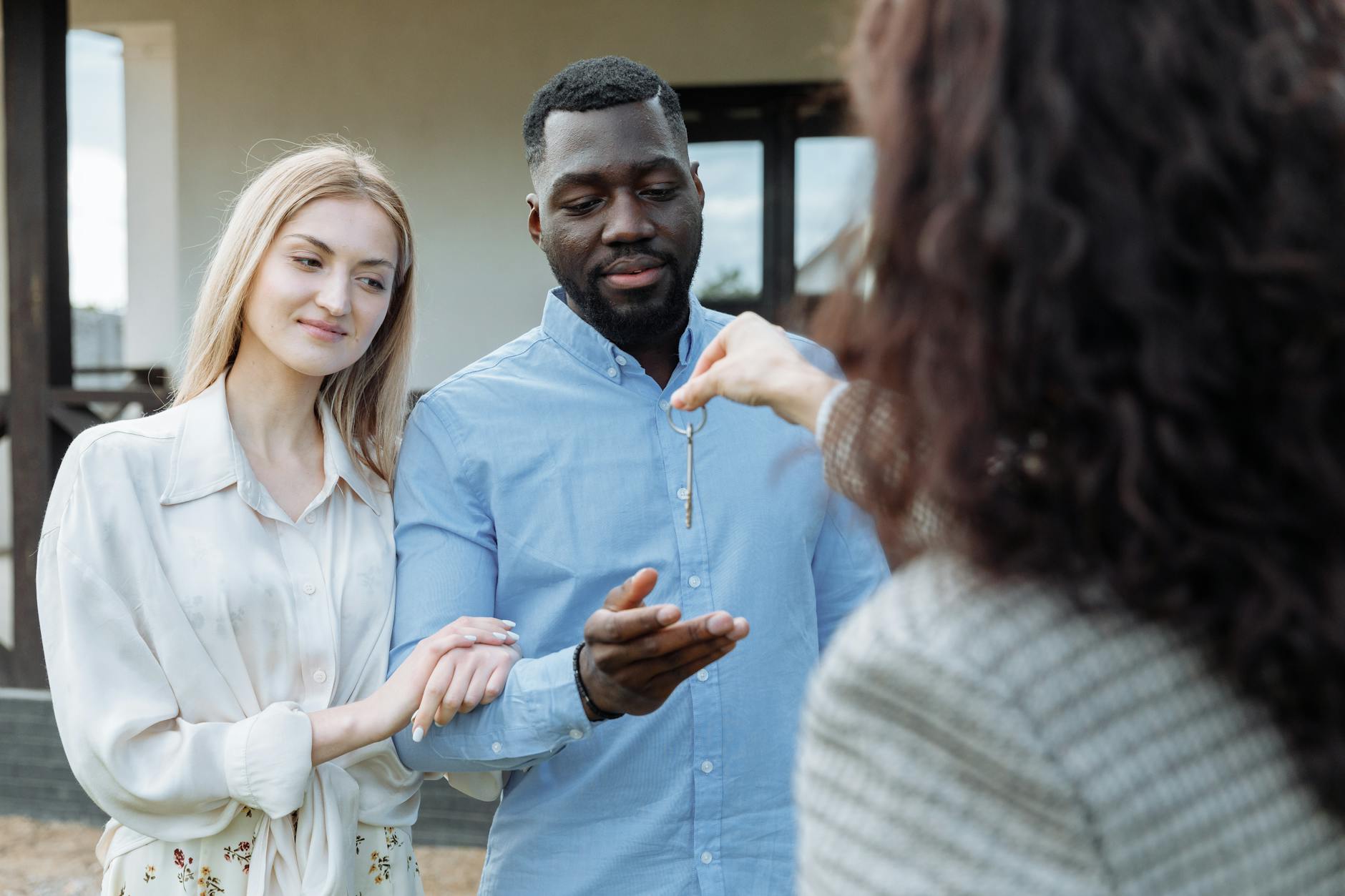A couple receive house keys from an estate agent outside a white rendered property on a sunny day