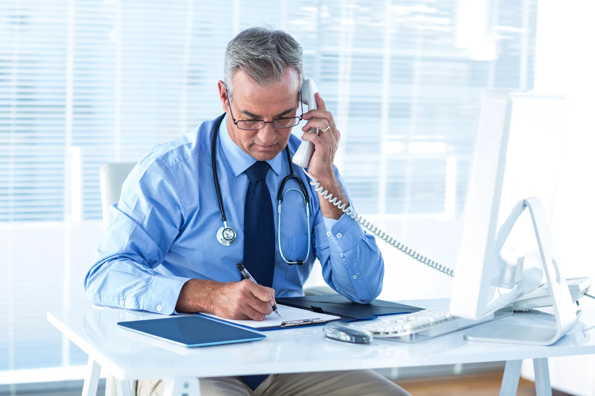 Middle-aged male physician in blue shirt and stethoscope taking notes while on a phone call at his desk in a modern high-rise office.