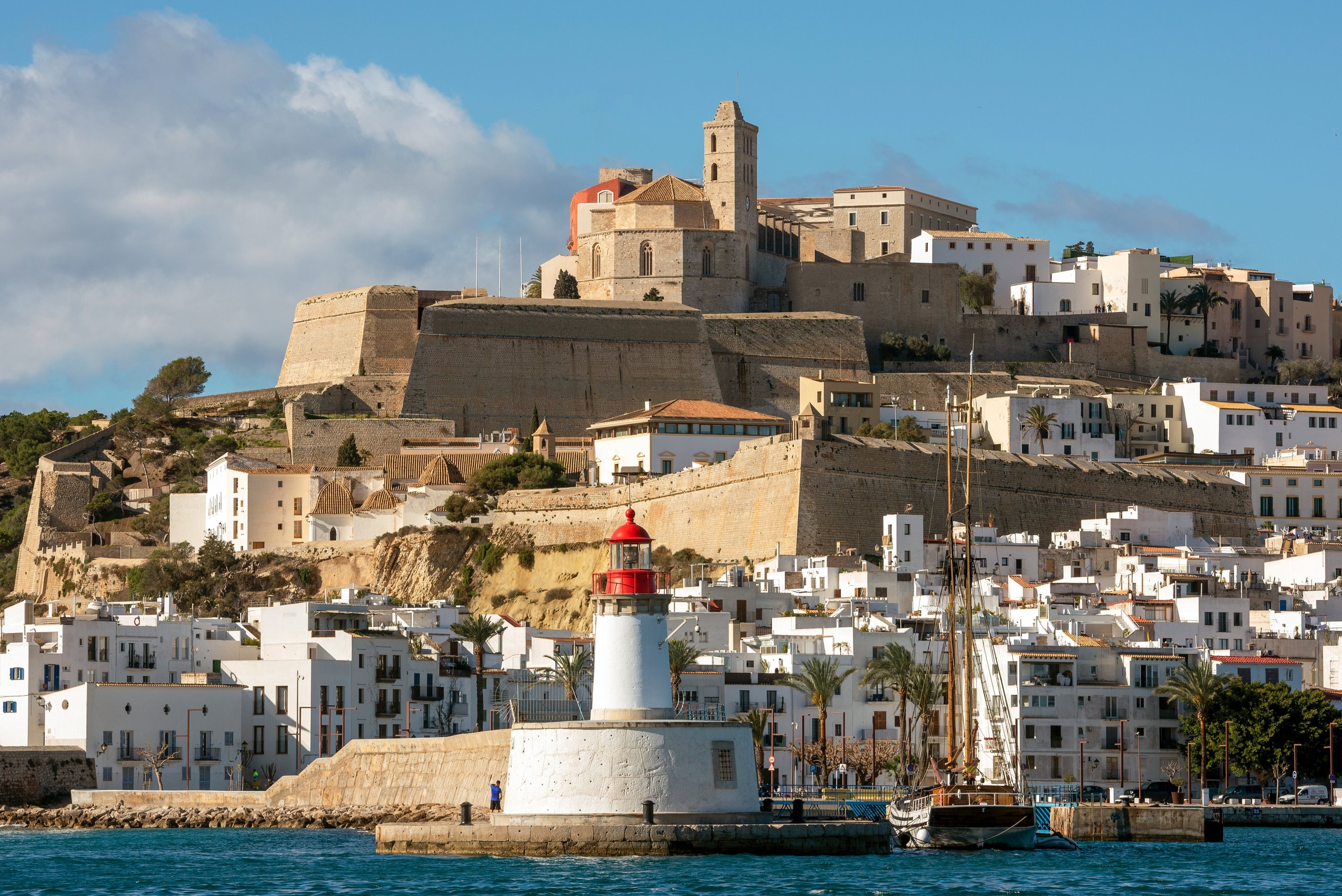 Dalt Vila — Ibiza's UNESCO-listed walled fortress old town, the cathedral and white houses descending the hill above the harbour, a wooden tall-ship sailboat moored below the lighthouse.