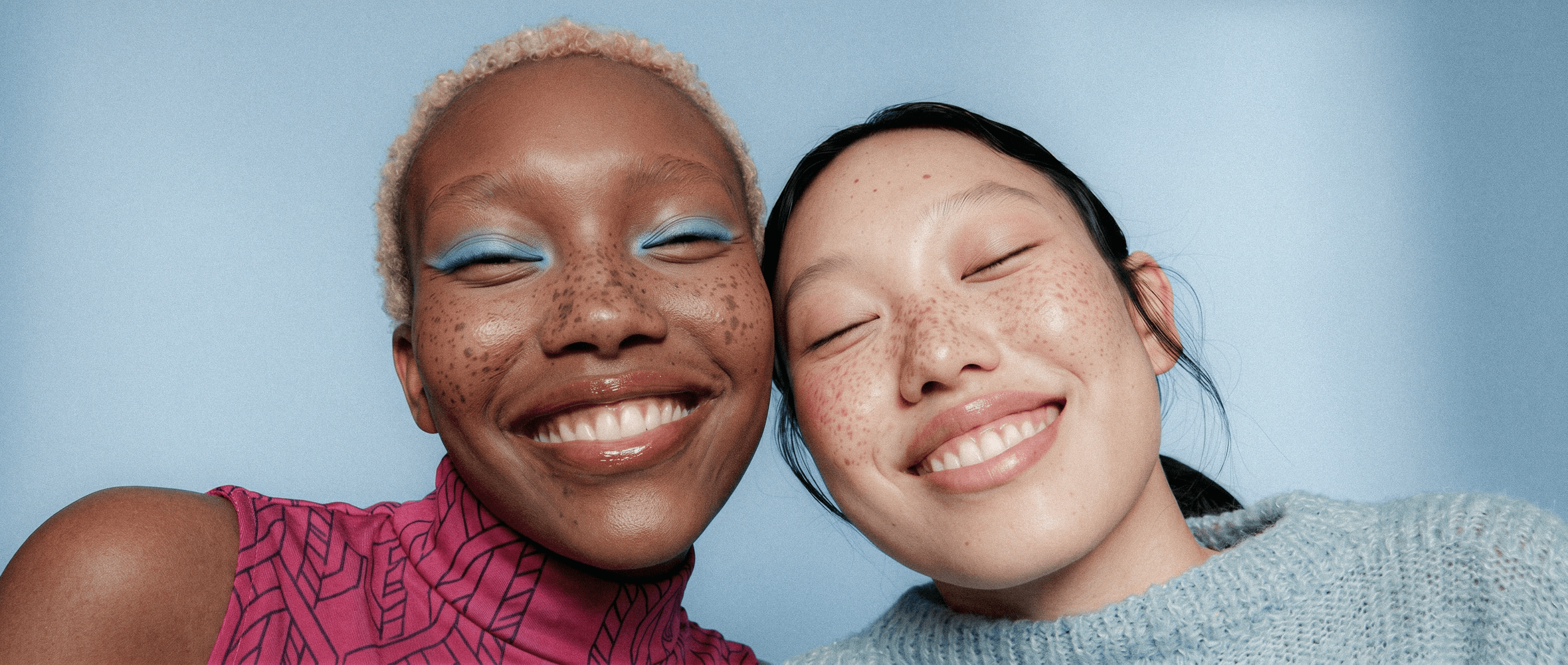 Close-up of two smiling friends with freckles, bright makeup, on a light blue background—joyful, inclusive brand portrait.