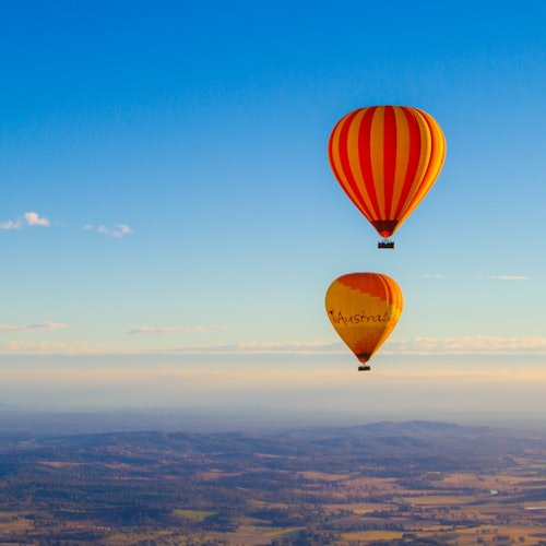 Twee heteluchtballonnen, één oranje met rode strepen en een andere oranje met 'Australië' erop geschreven, zweven boven een schilderachtig landschap.