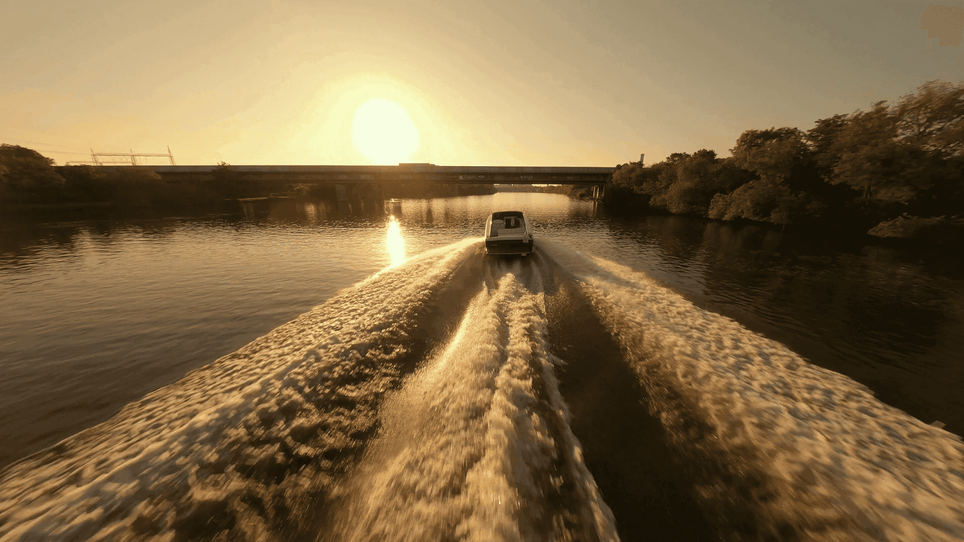 A white van drives across the water, leaving a frothy wake under a golden sunset, with a silhouette of a bridge and lush trees on the horizon.