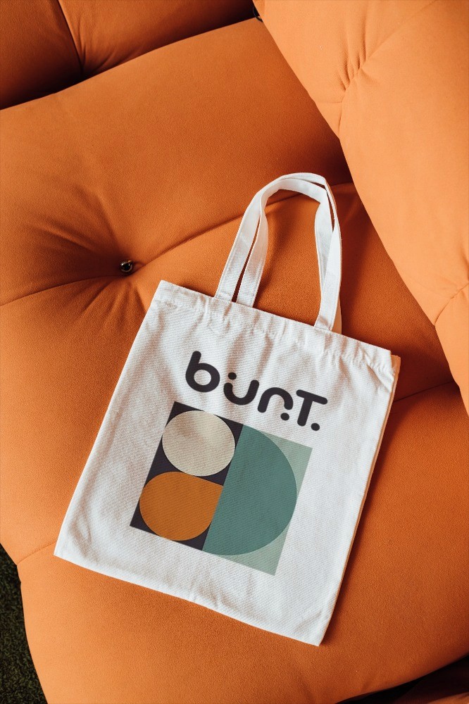 Beiger Baumwoll-Beutel mit mehrfarbigem bunt.ist Logo-Print in Orange, Grün und Rosa auf terrakotta-orangefarbenem Hintergrund