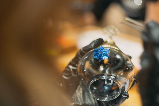 A person crafting a Turkish mosaic lamp, carefully applying multicolored beads to the ornament. The background shows a workshop setting with tools hanging on the wall.