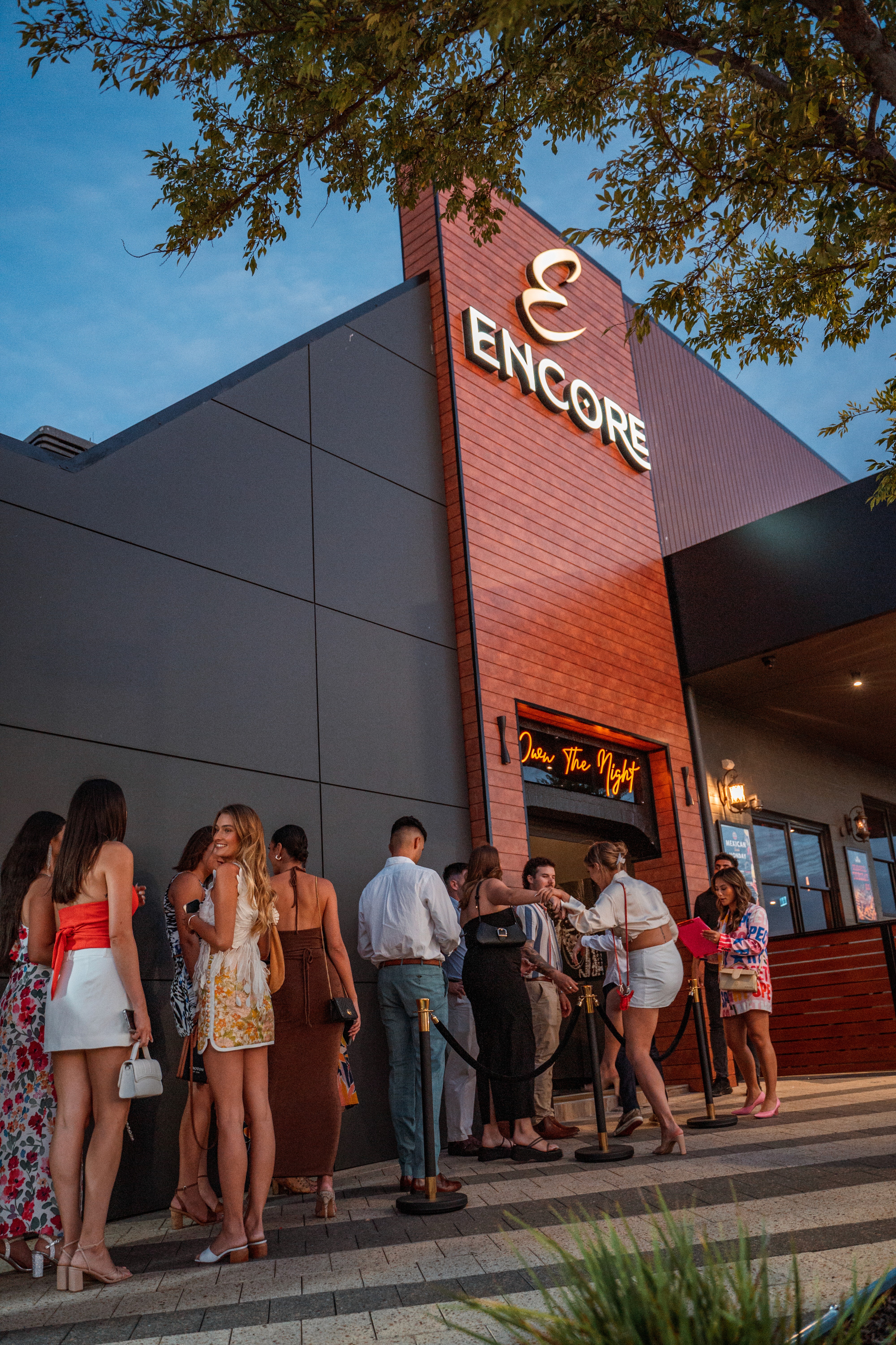 Exterior view of Encore Rockingham at night, featuring a 'Join the Night' neon sign and guests lined up behind velvet ropes.