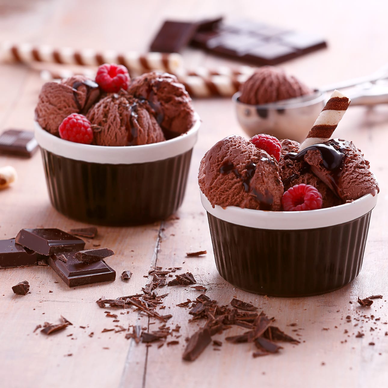 Dwie miski czekoladowych lodów z malinami, sosem, wiórkami i rurkami na drewnianym stole