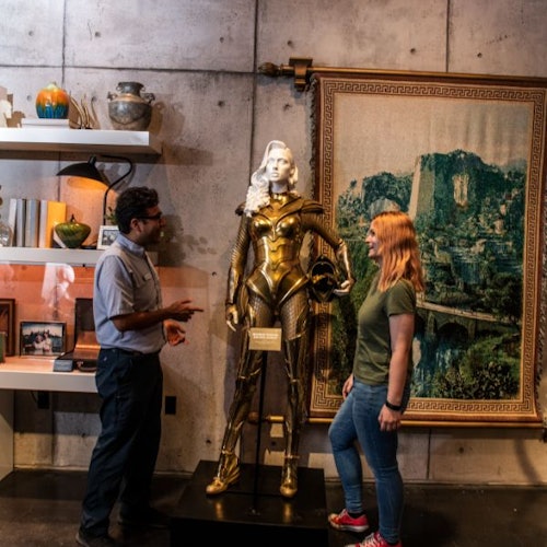 Duas pessoas estão ao lado de uma estátua de guerreiro dourado em uma sala com prateleiras de decoração e uma tapeçaria na parede.
