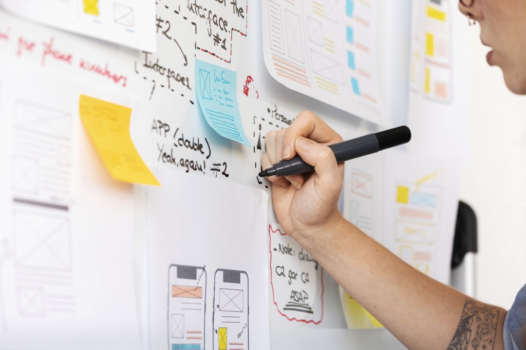 Visual of A person sketches app wireframes and notes on a whiteboard covered with sticky notes and design drafts.