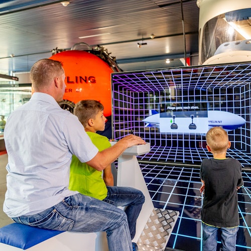 A man and two boys interact with a digital exhibit showcasing a grid and diagrams, with a large orange object in the background.