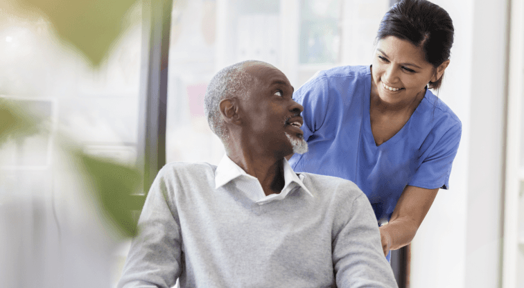 A smiling elderly man receiving attentive care from a nurse in blue scrubs in a care or home setting