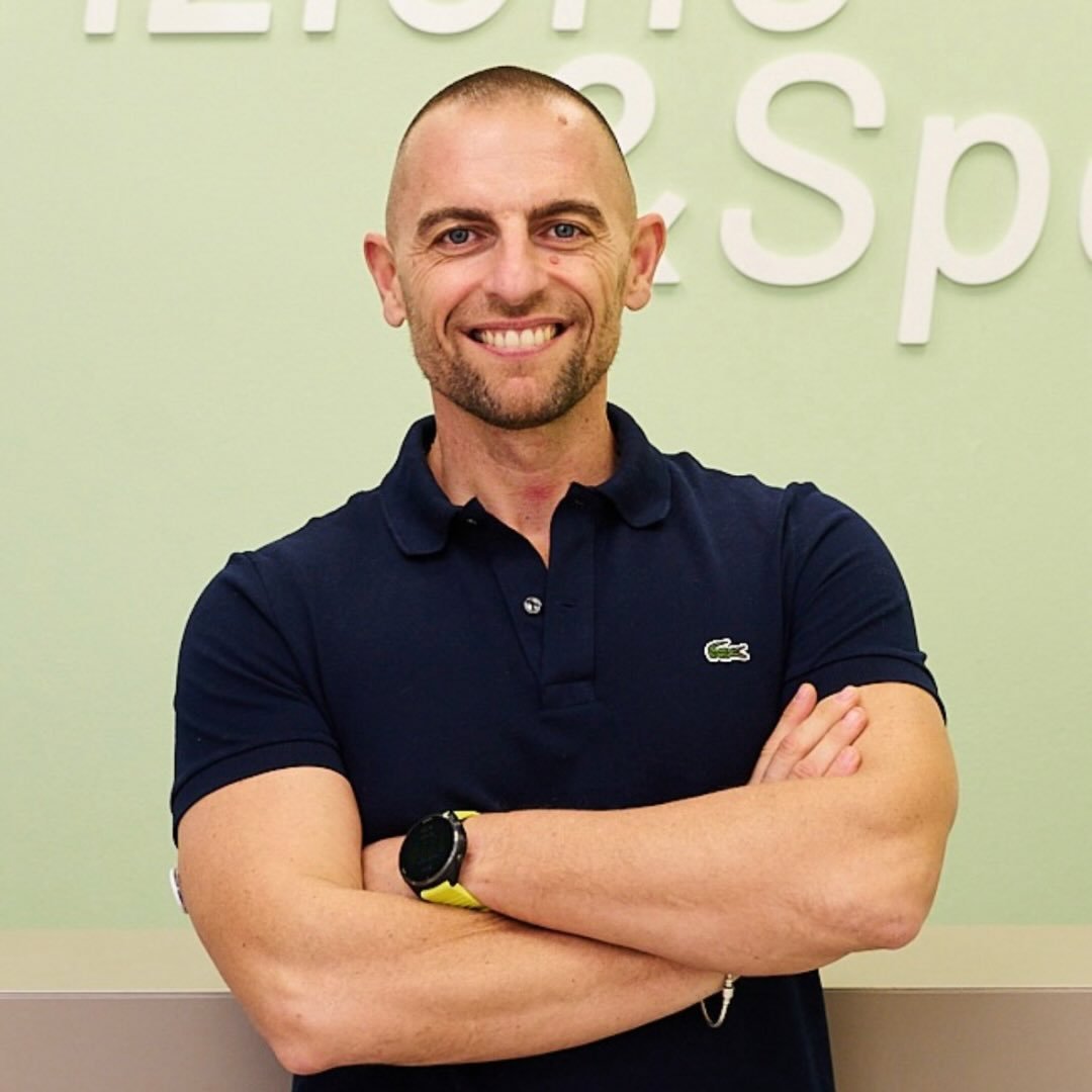 A woman with blonde bob hair in a grey coat and beige turtleneck stands against a neutral backdrop. She has a serious expression, conveying elegance.