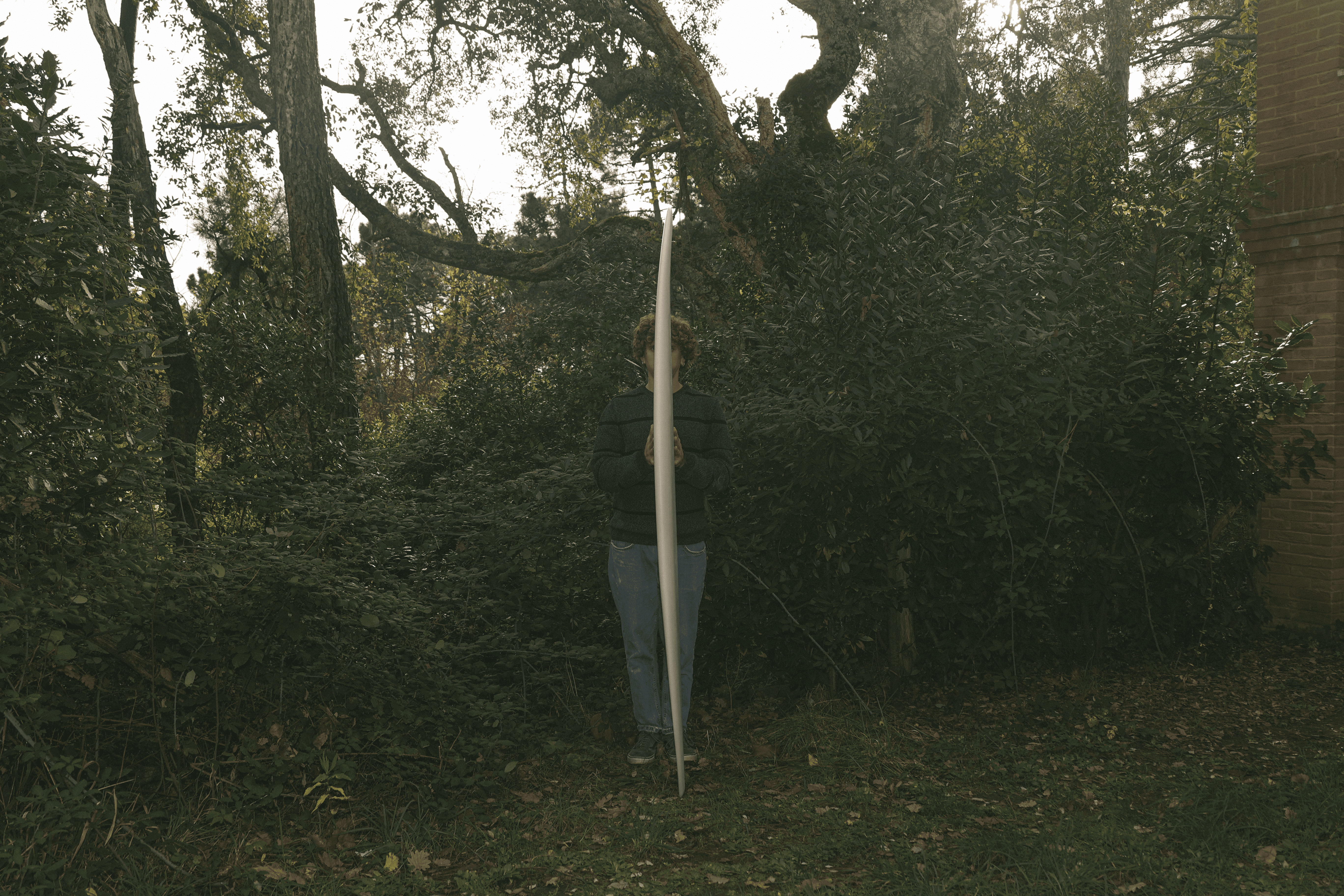 Una persona in piedi in un ambiente boschivo tiene in verticale una tavola da surf stretta e bianca che copre la parte superiore del corpo.