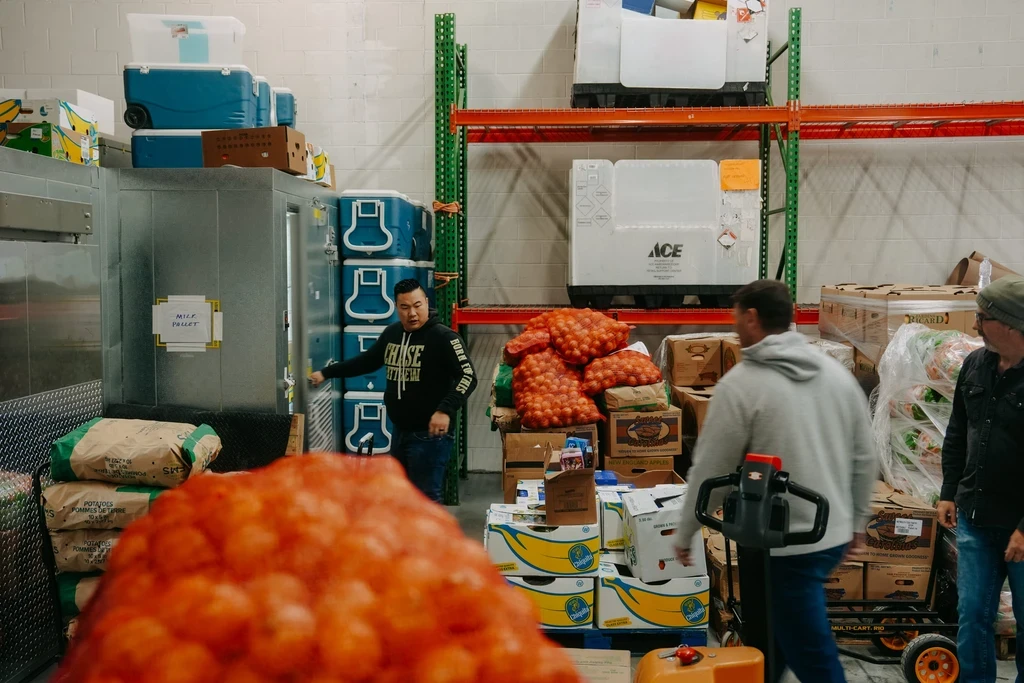 A photograph inside a warehouse or food distribution center. A man wearing a dark sweatshirt with white text is opening the silver door of a walk-in refrigerator unit on the left. The scene is busy with stacked inventory: large mesh bags of orange produce (onions or oranges) are piled on a pallet in the center. High orange metal shelves are visible in the background, one with a large white unit labeled "ACE". Two other men are partially visible on the right, one operating a pallet jack in the foreground.