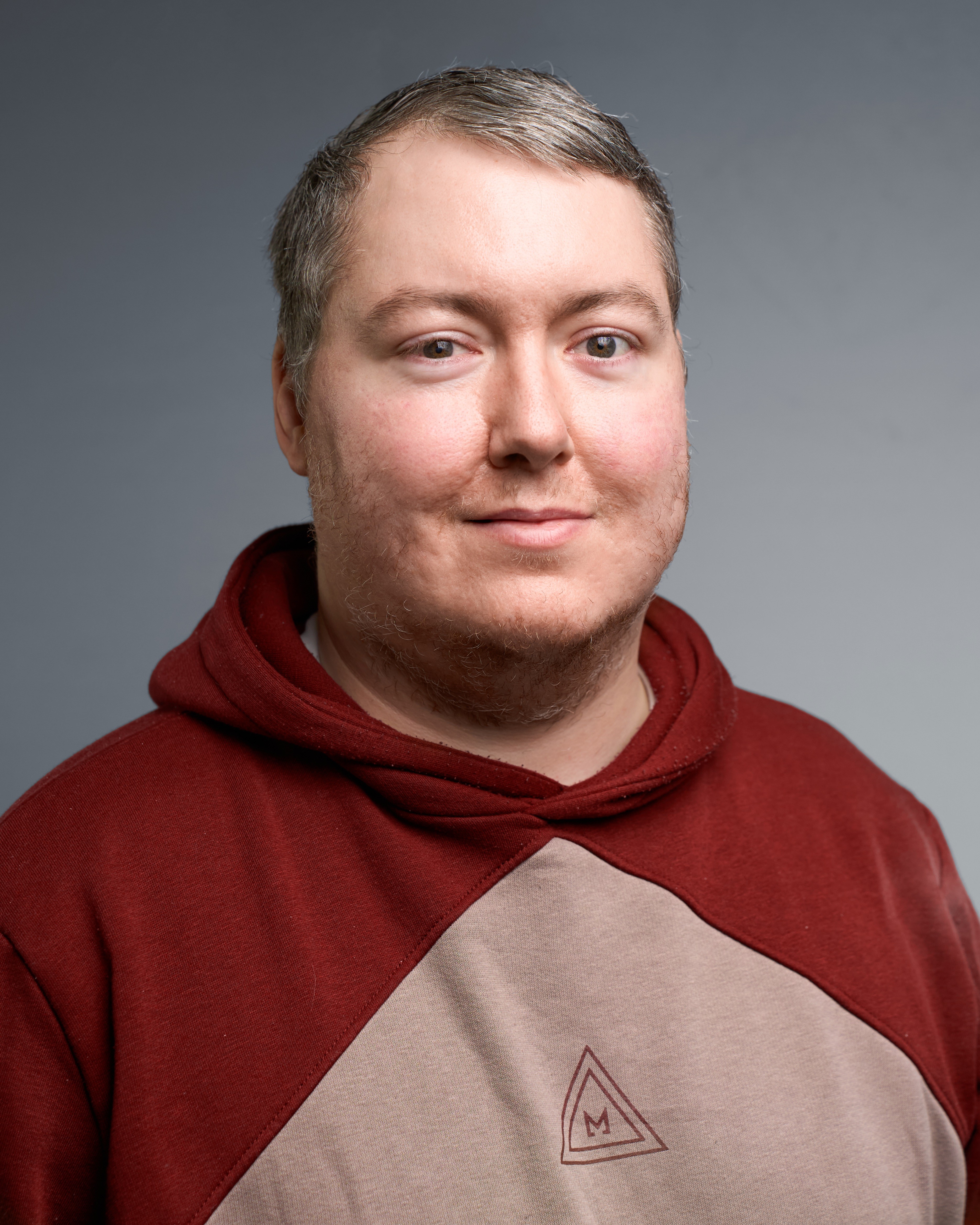 An image of Mark Maddison, wearing a red and light pink hoodie, on a grey background. Mark has short, grey hair. He is smiling.