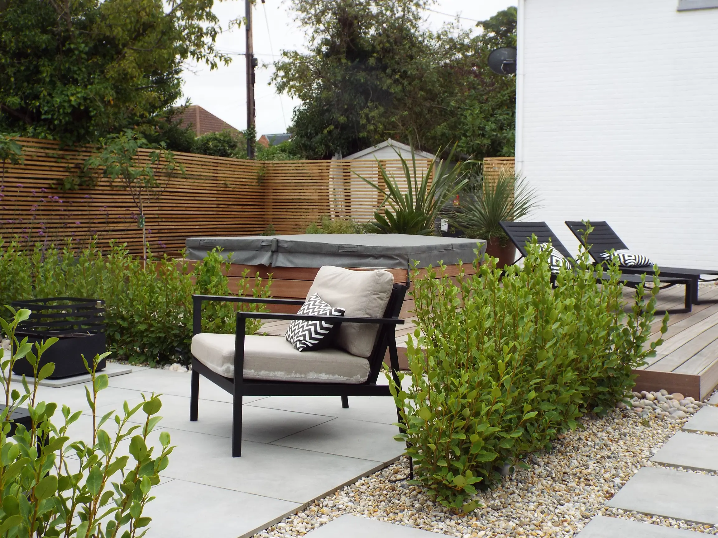 A serene garden area featuring a lounge chair, greenery, and a paved pathway beside a white wall.