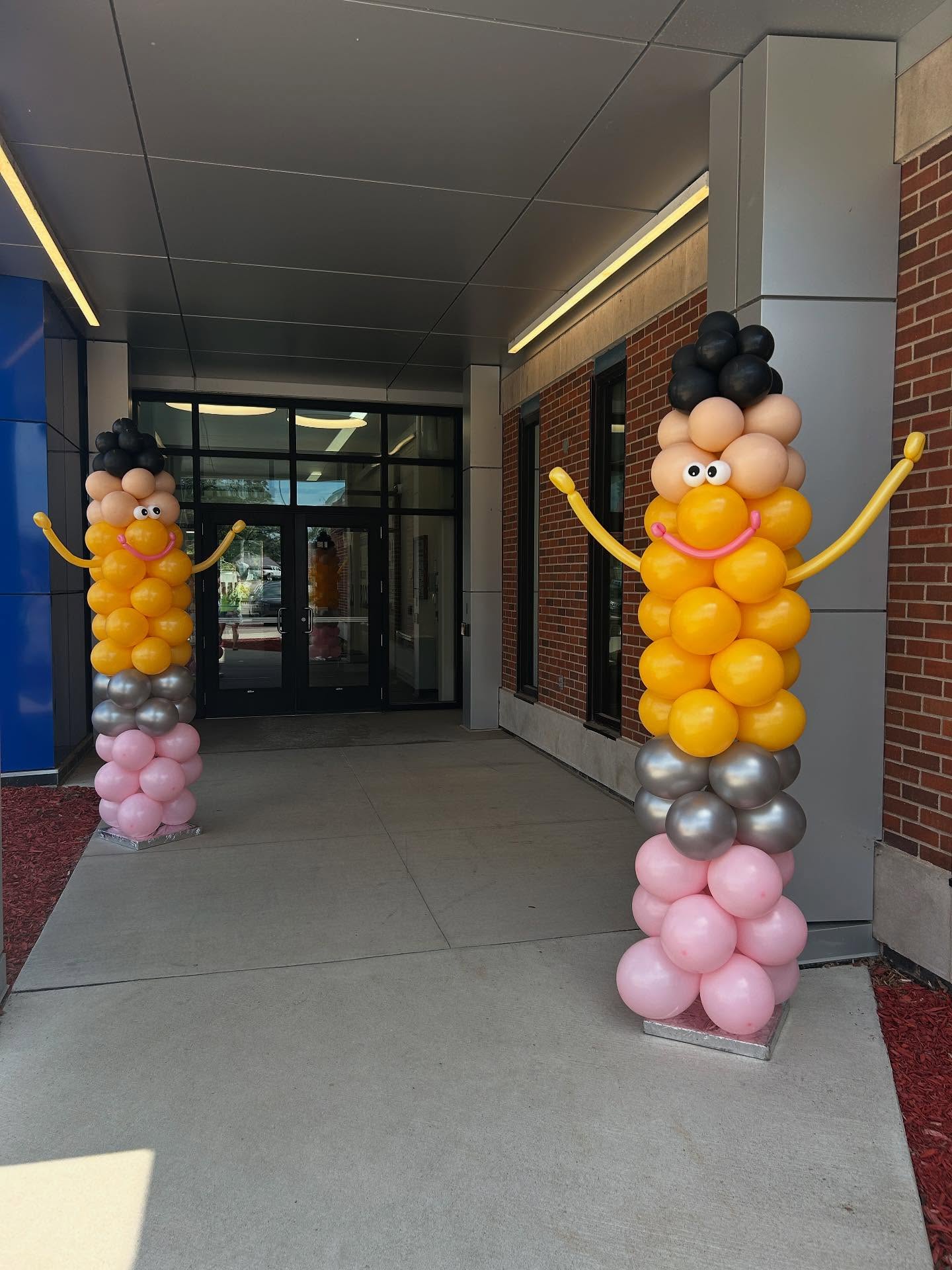 Two tall balloon columns resembling cheerful figures with faces and outstretched arms stand at the entrance of a building, made with yellow, black, gray, and pink balloons.