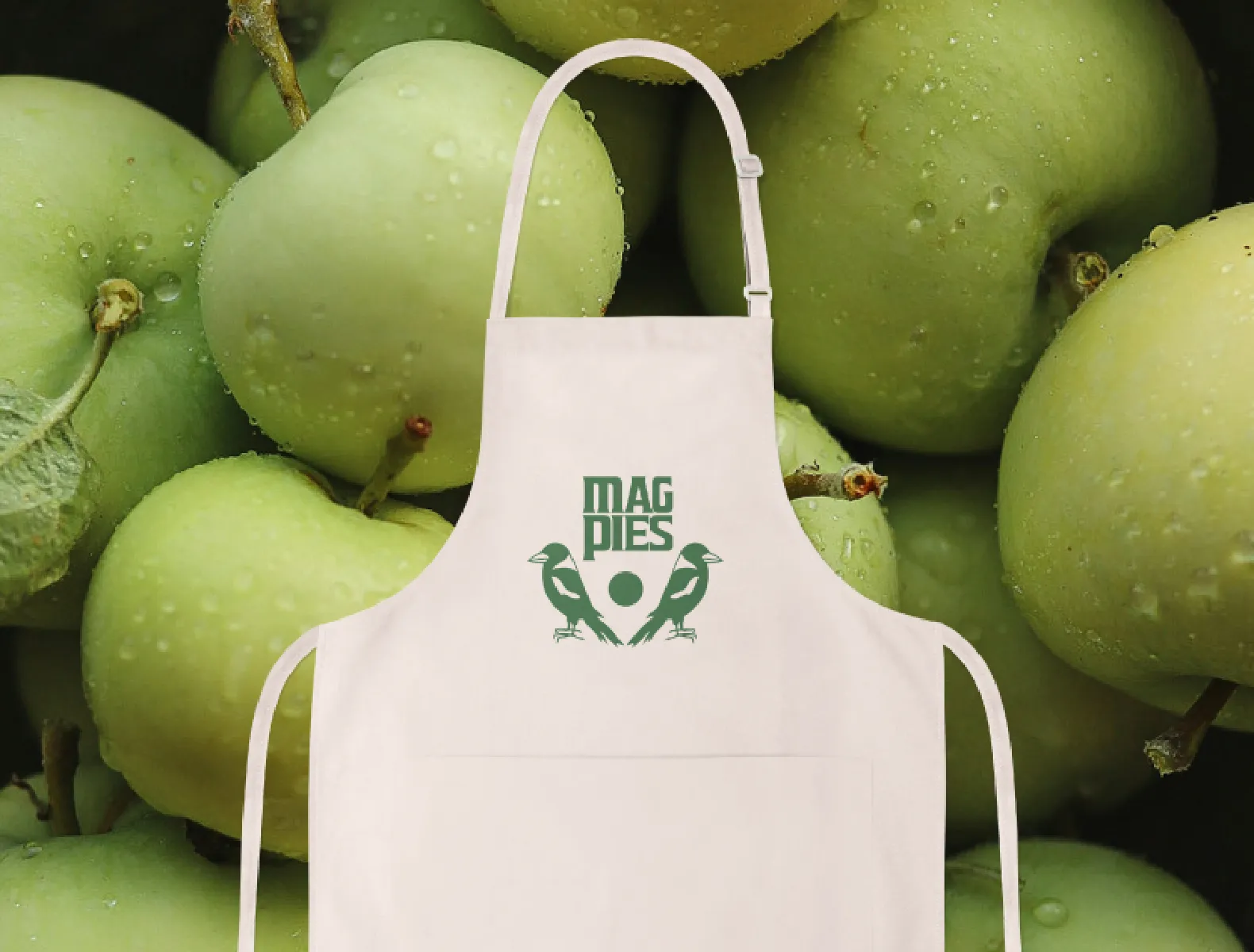 The primary MagPies logo printed in green on a canvas apron with granny smith apples in the background.