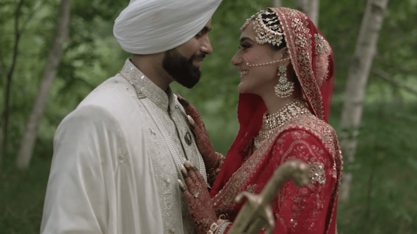 Man in off-white attire and woman in red smiling at each other, with the woman’s hands placed on the man’s chest in a loving moment