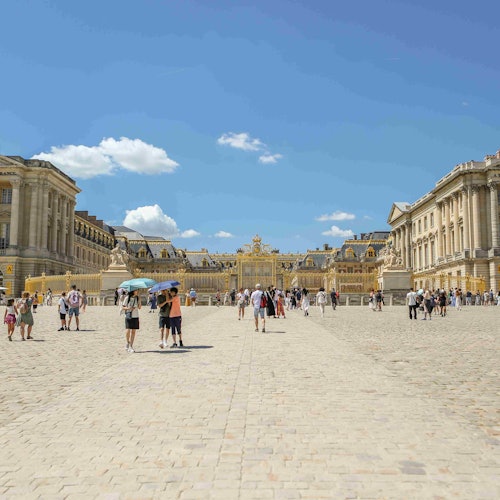 Personas caminando y tomando fotos en un gran patio de adoquines con majestuosos edificios y una elaborada puerta dorada bajo un cielo azul.