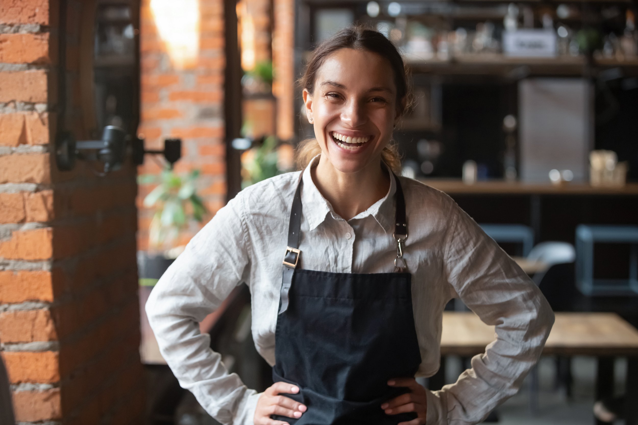 A smiling woman in a white shirt and black apron standing inside a cafe or restaurant.