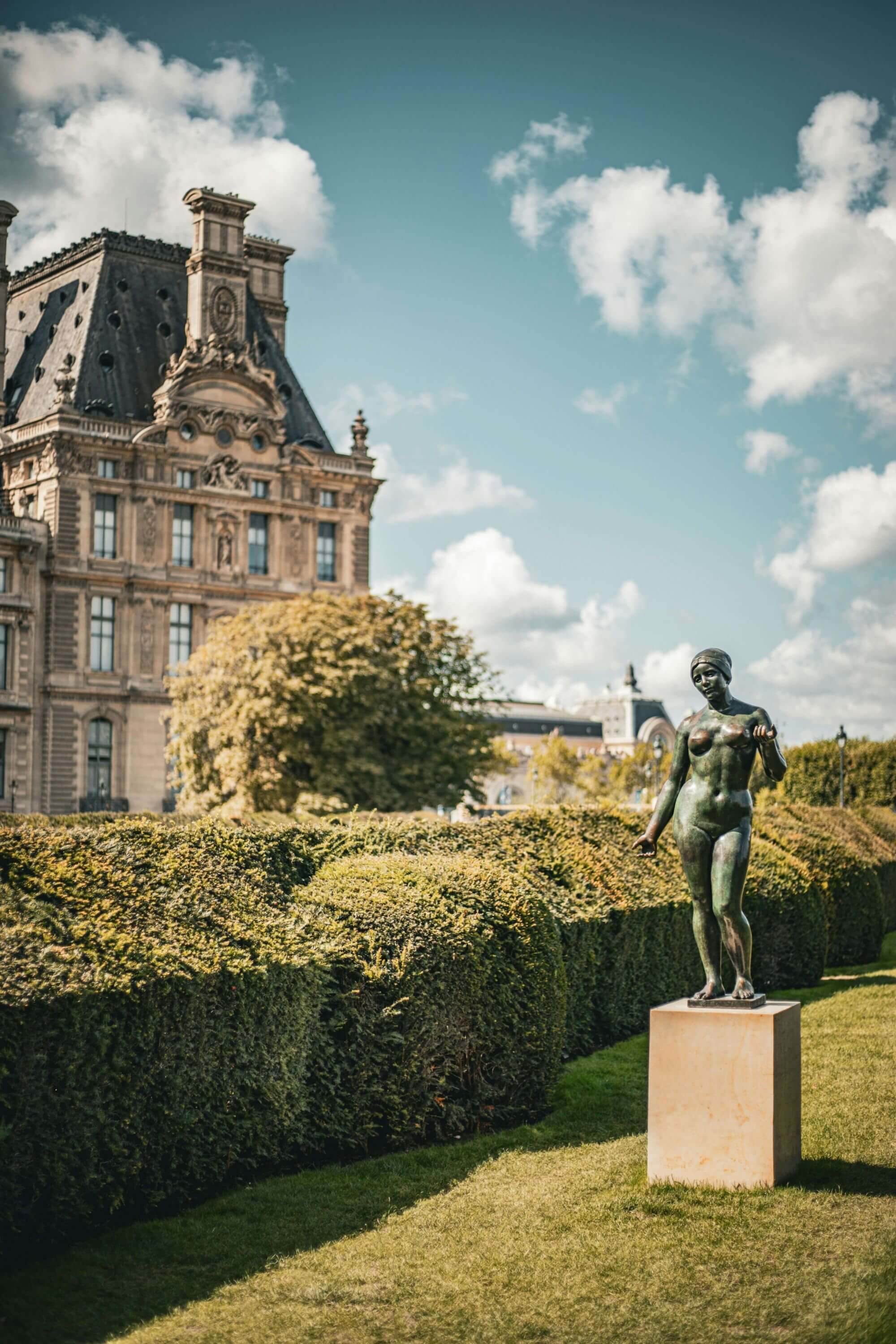 Historic stone building with a tall roof next to a lush garden. A bronze statue stands on a pedestal, under a partly cloudy sky. Serene ambiance.