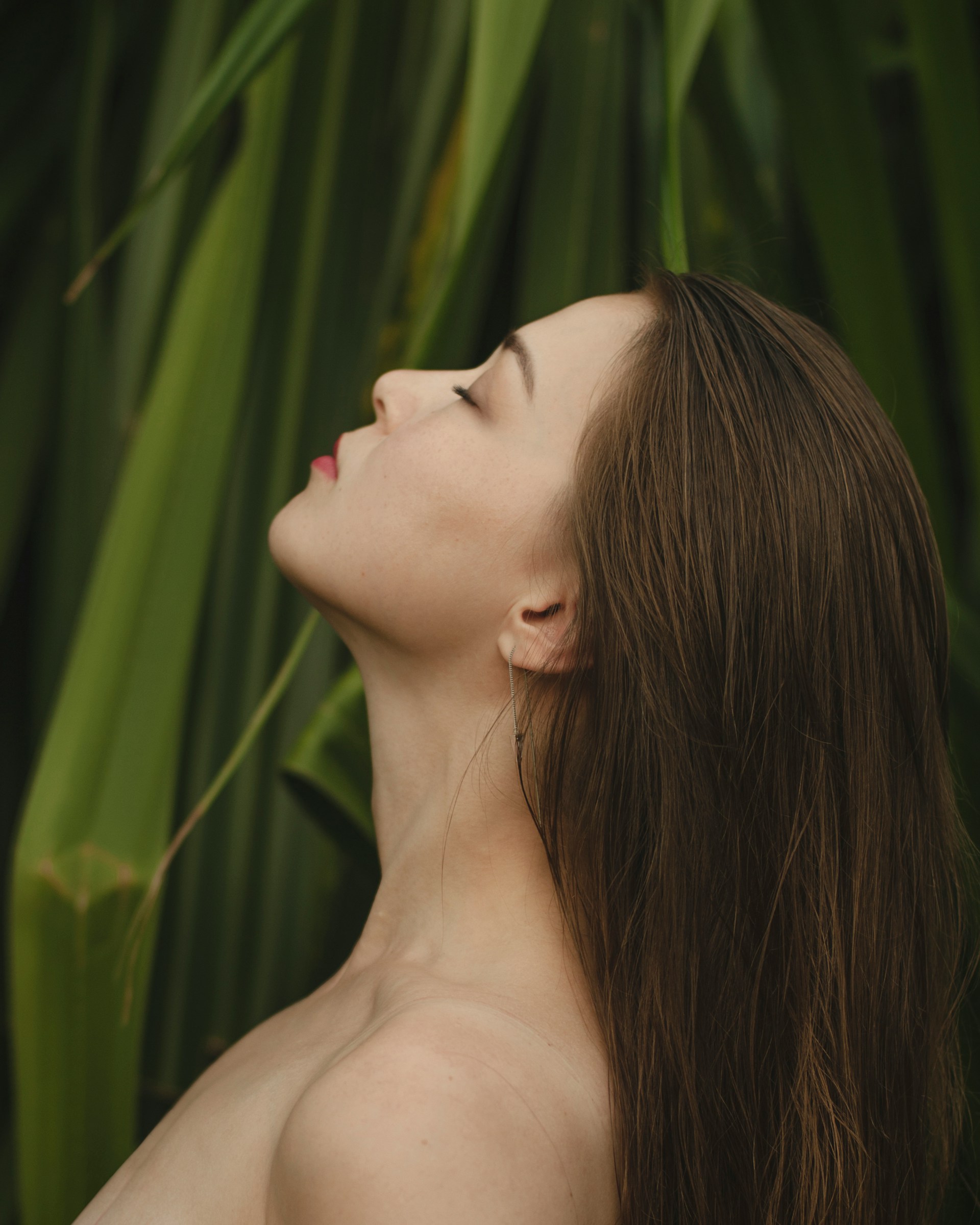 A side profile of a woman with long brown hair and her eyes closed, posed against a backdrop of large, green tropical leaves.