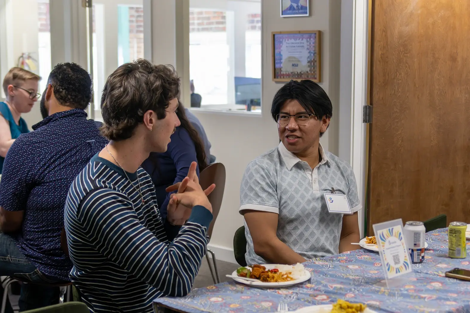A group of people sits around a table covered with a patterned tablecloth, engaging in conversation while enjoying a meal, with one person gesturing expressively to another.