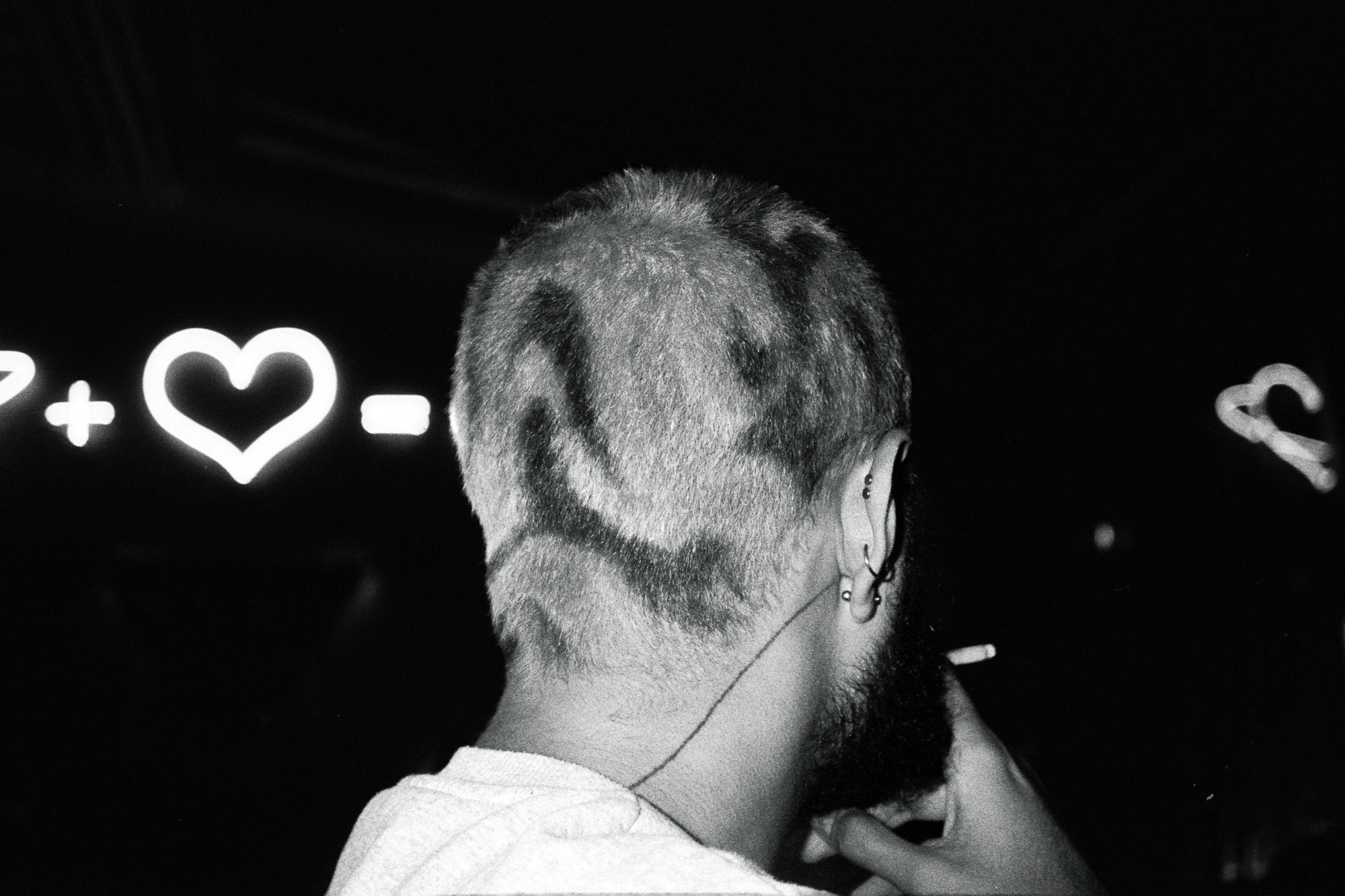 A person with short, intricately shaved hair featuring abstract patterns enjoys a smoke in a dimly lit area, with glowing mathematical symbols and a heart illuminated in the background.