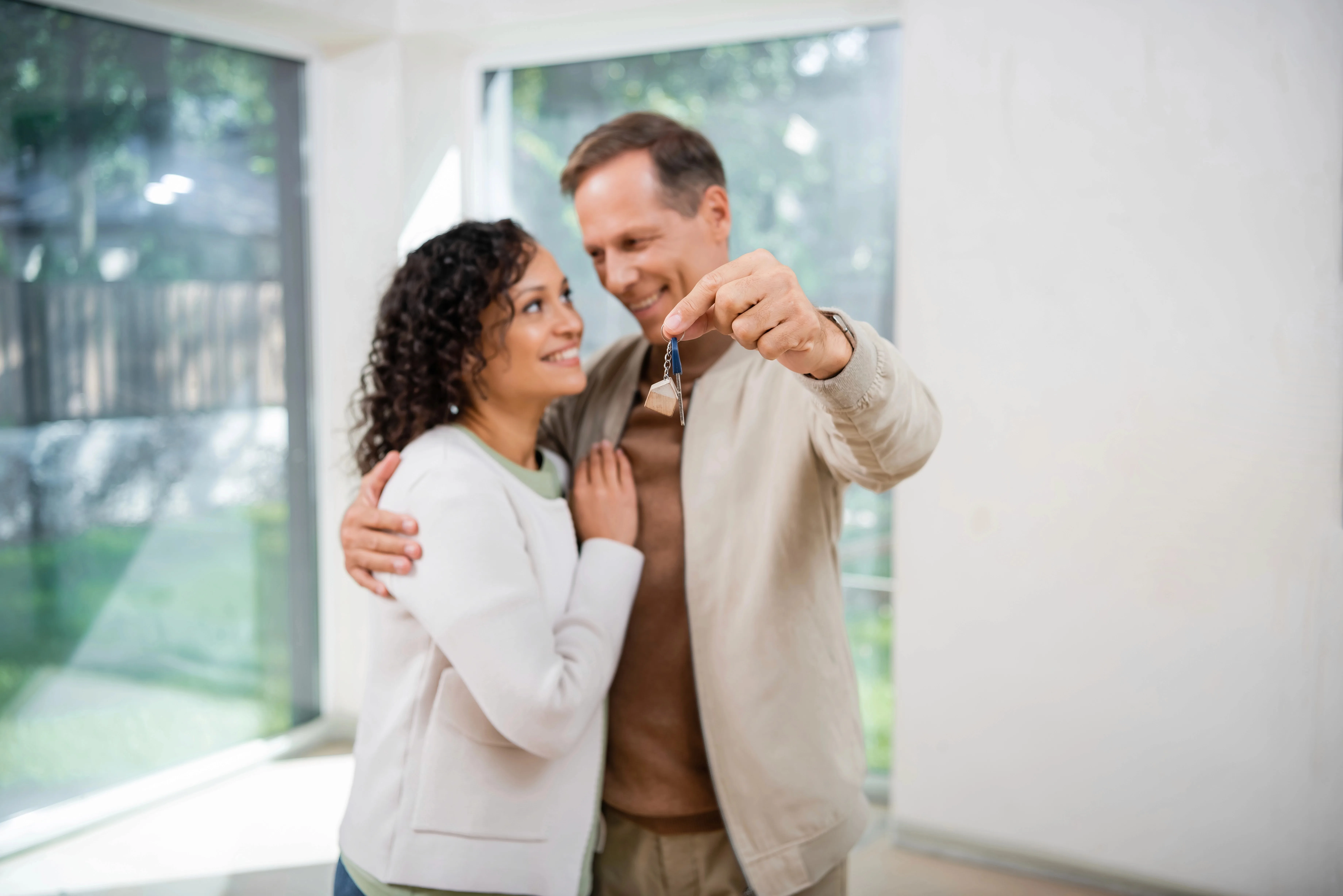 A couple stands together in a bright, modern home with large windows, smiling as the man holds up a house key.