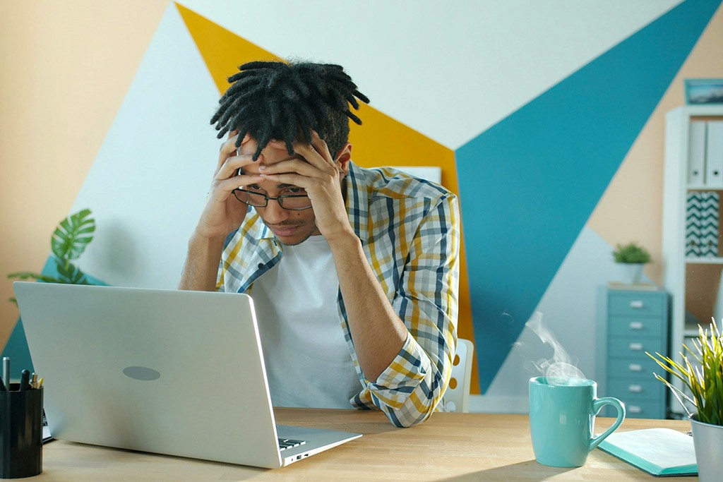 Un homme stressé regarde son ordinateur portable, la tête entre les mains, assis à un bureau avec une tasse fumante.