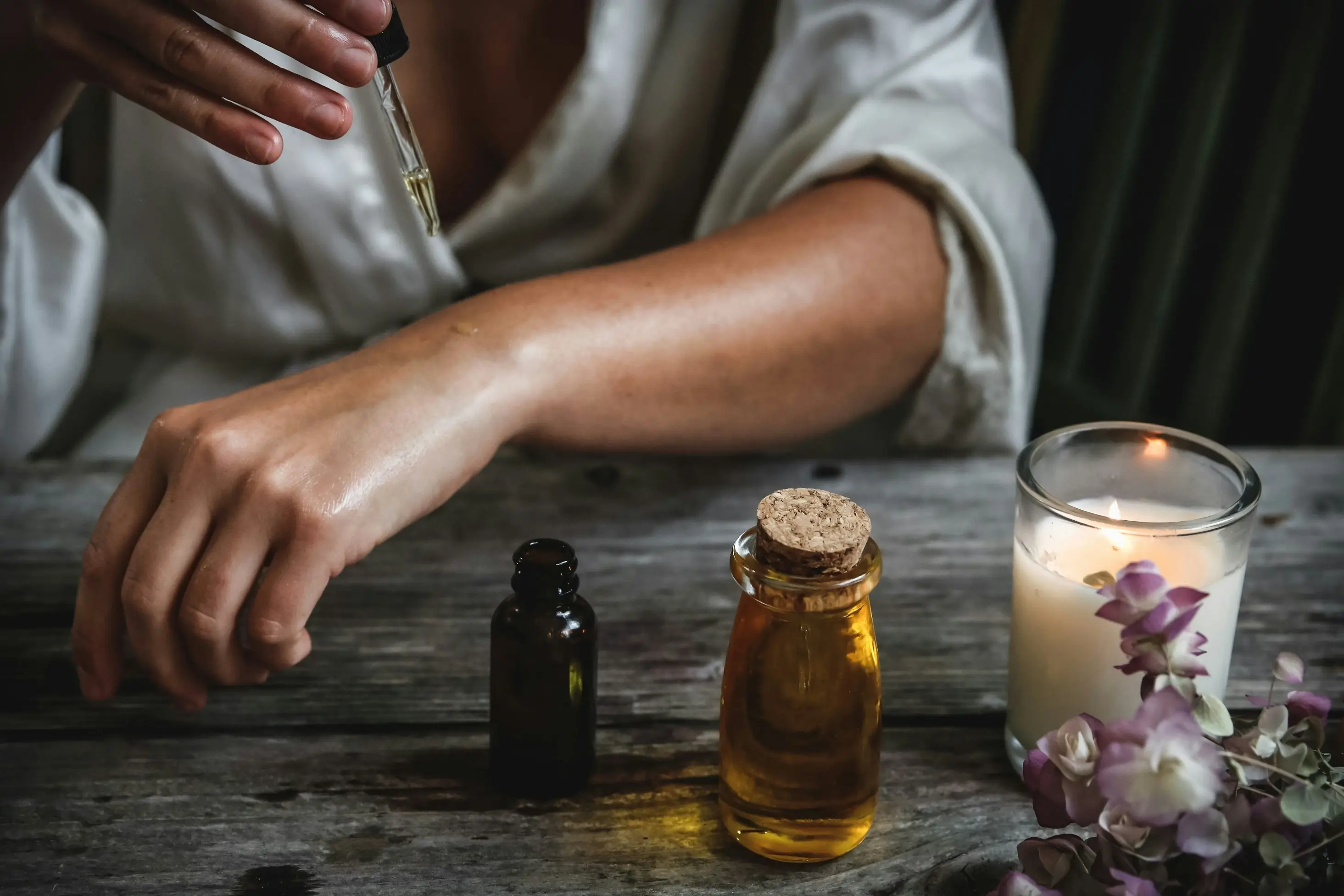 Person applying essential oil to their arm with a dropper, surrounded by a rustic setup featuring glass bottles, a lit candle, and fresh flowers on a wooden table.