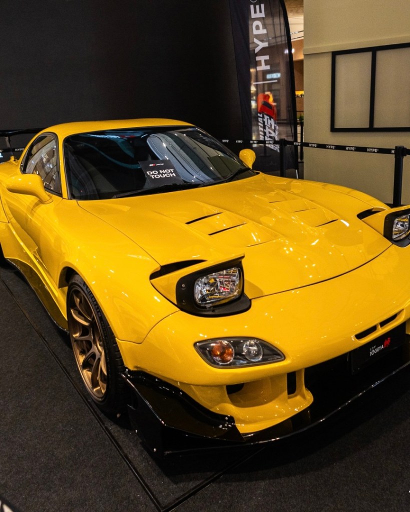 A yellow sports car on display at a pop-up event, featuring custom modifications and a 'DO NOT TOUCH' sign in the windshield.