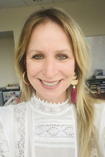 Close-up photo of a woman with blond hair smiling widely. She wears large gold hoop earrings with tassels and a white blouse.