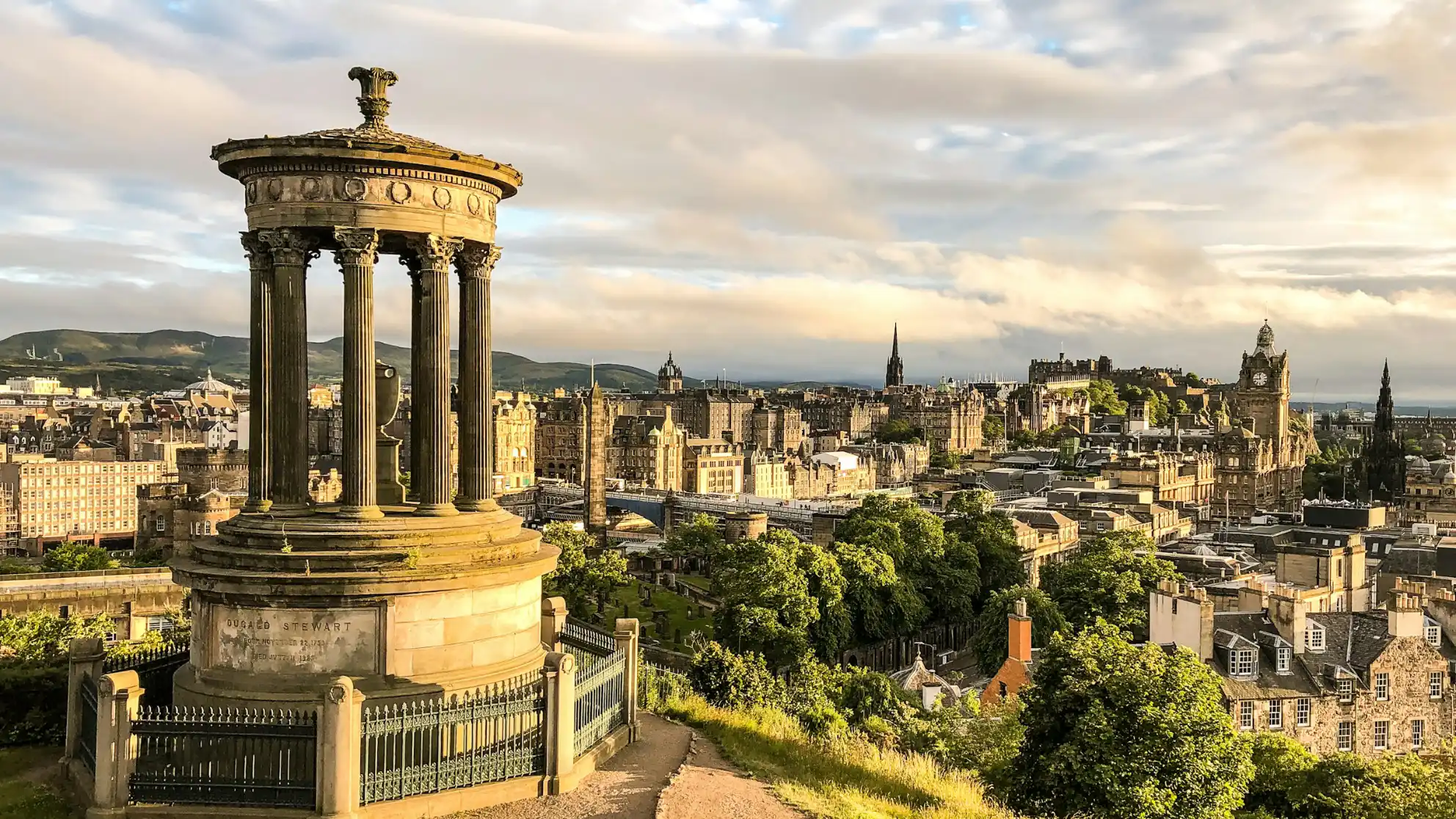 A neoclassical monument with columns stands in the foreground, overlooking Edinburgh's historic skyline under a golden, late afternoon sky.