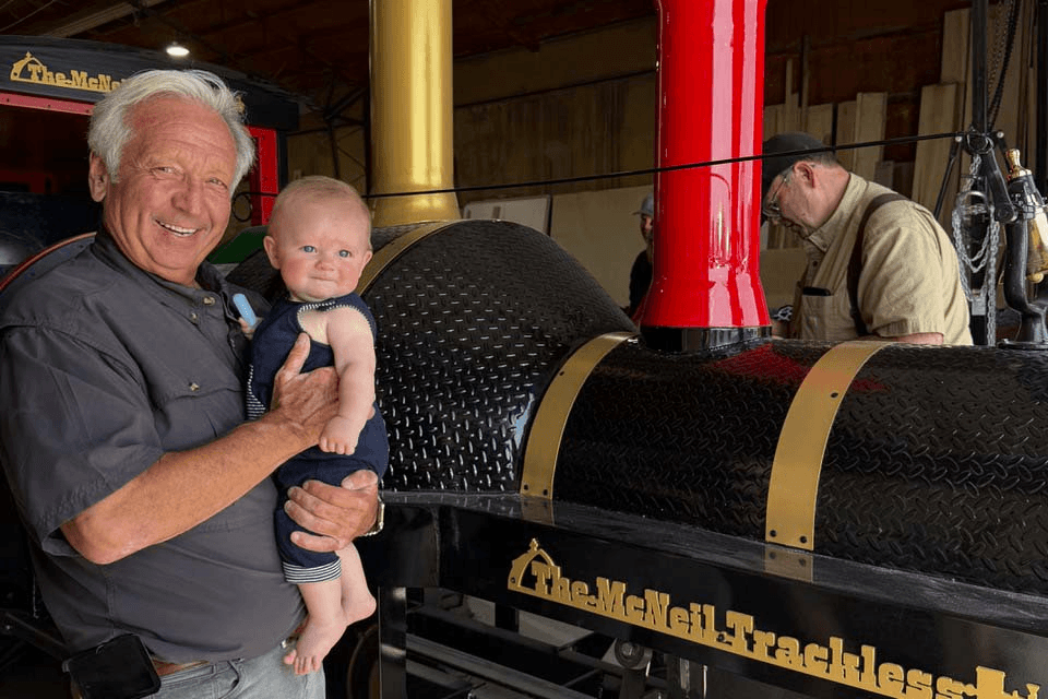 Rollie Walker holding a grand child in front of McNeil Trackless in shop