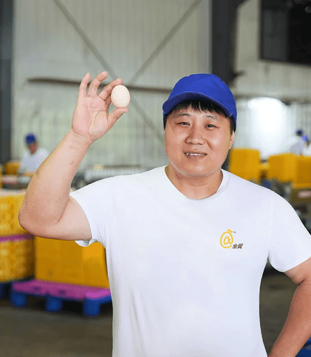 A smiling Jinyi Food worker in a blue cap proudly displays a single fresh egg, symbolizing quality egg products.