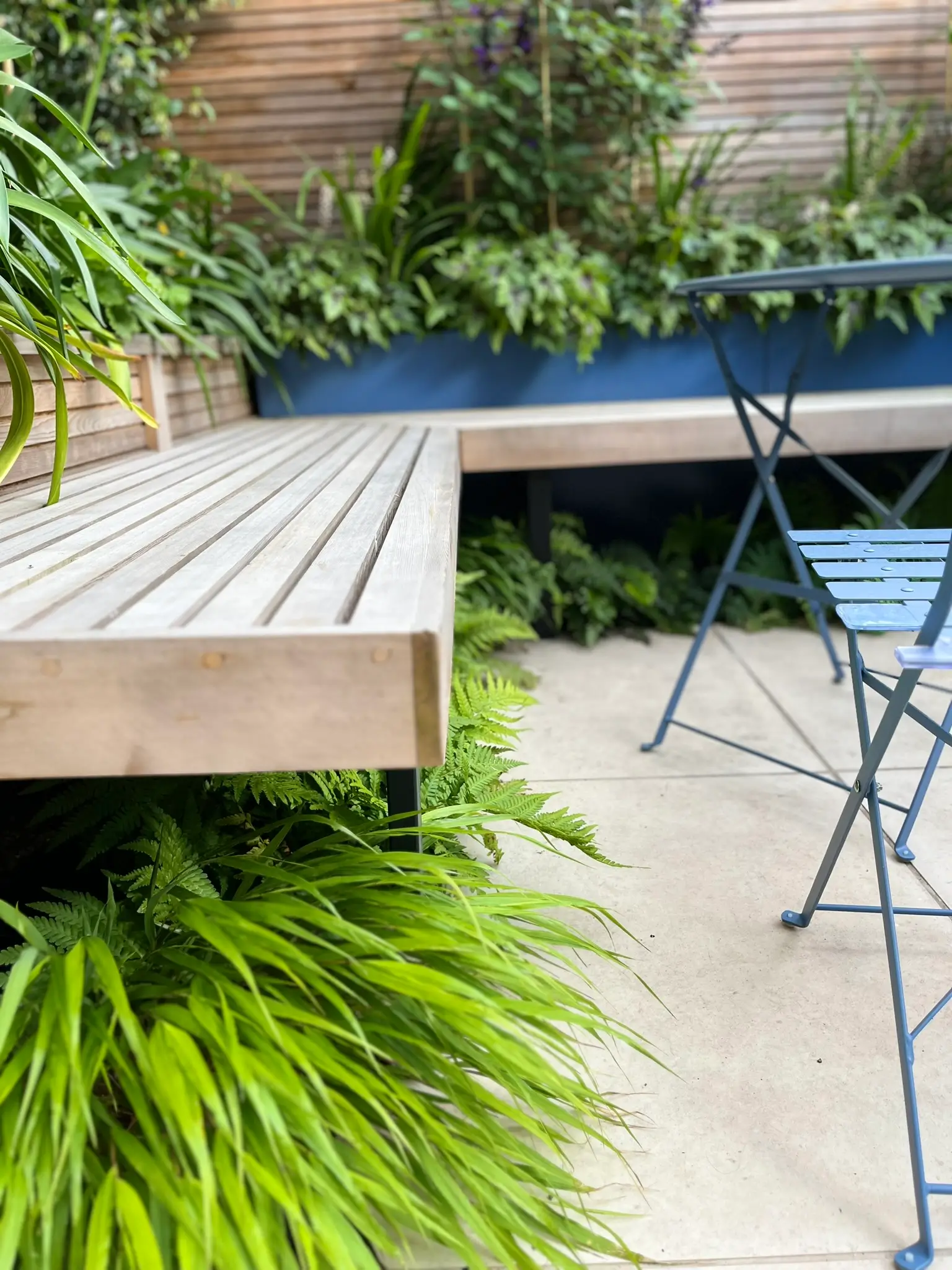A wooden deck blends into a garden, surrounded by lush greenery and a blue railing. A chair is nearby.