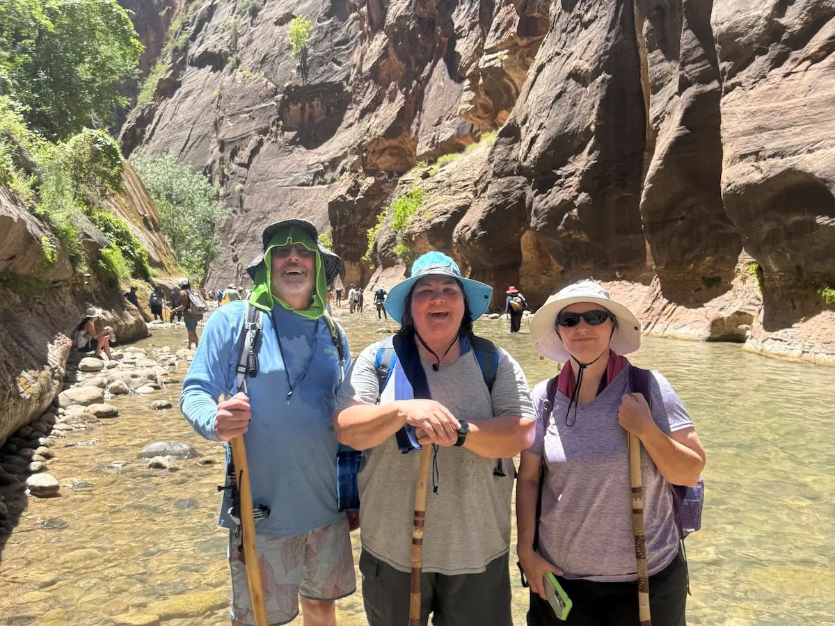 Stu, his sister Sara, and their friend Carrie standing in the Narrows river in Zion National Park, wet from the hike and holding walking sticks during a summer trip.