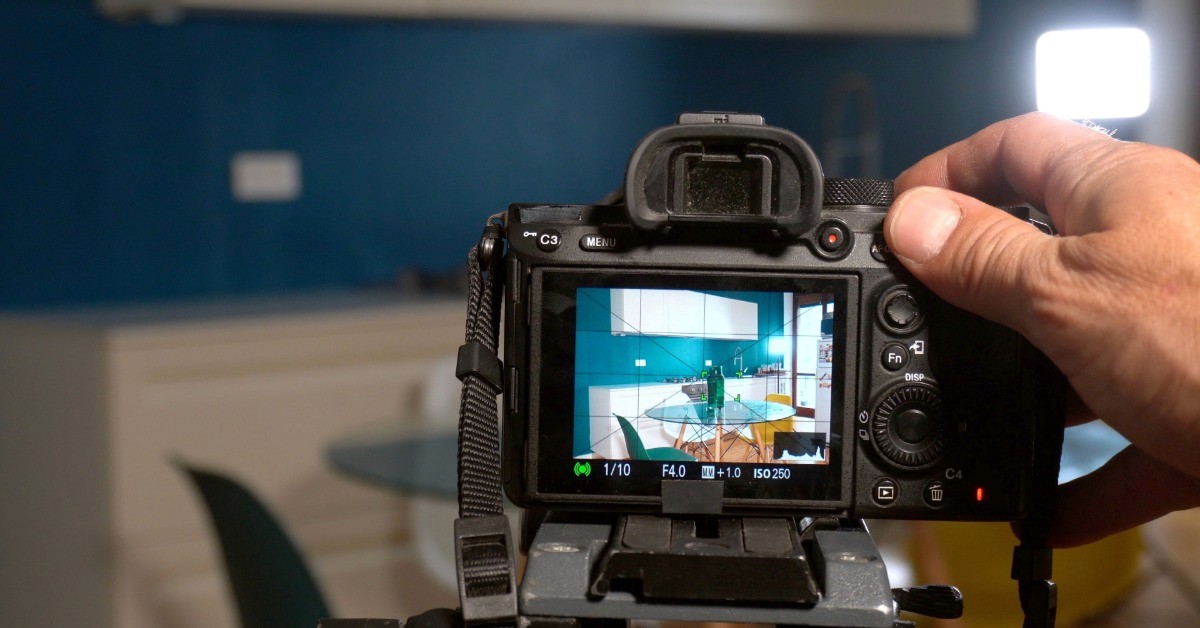 A close-up of a camera set up on a tripod. The camera screen shows a dining room table with white cabinets in the background.