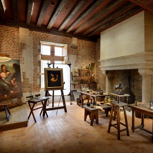 A historical artist's studio with wooden tables, painting easels, art supplies, and a large fireplace.