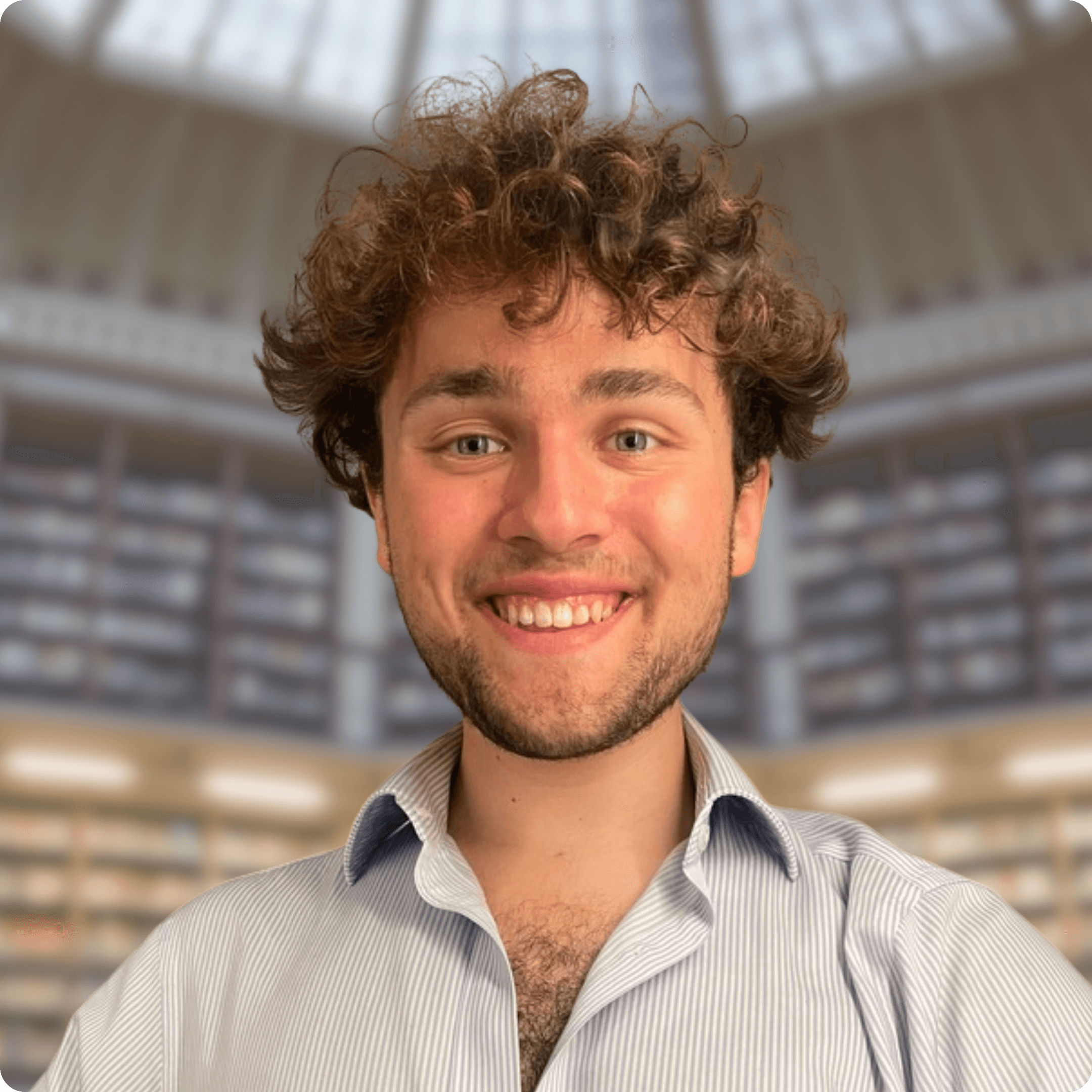 A young person with curly hair smiles at the camera while sitting in front of a laptop against a dark background.