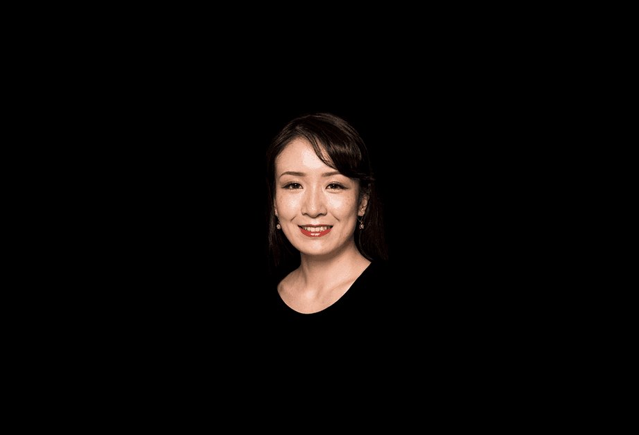 A woman with dark hair pulled back is centered against a solid black background. She is wearing a black top and small, light-colored earrings. Her face is lit with a warm smile, revealing her teeth. Her eyes are open and looking directly at the camera. The lighting is focused on her face, leaving the background dark and undefined.