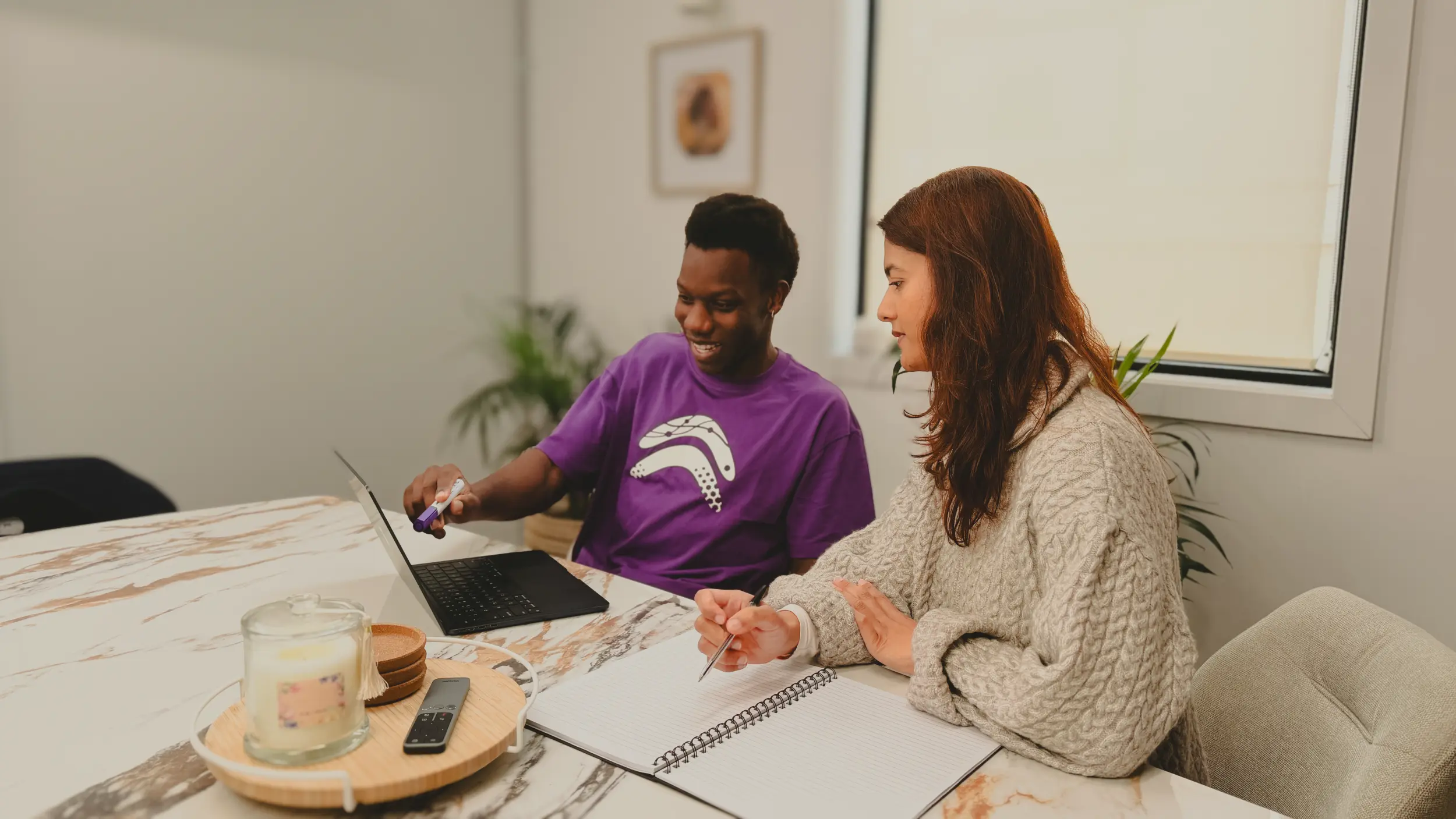 Two colleagues reviewing work together at a desk with a laptop and notebook,  smiling and engaged in a collaborative recruitment workflow session.