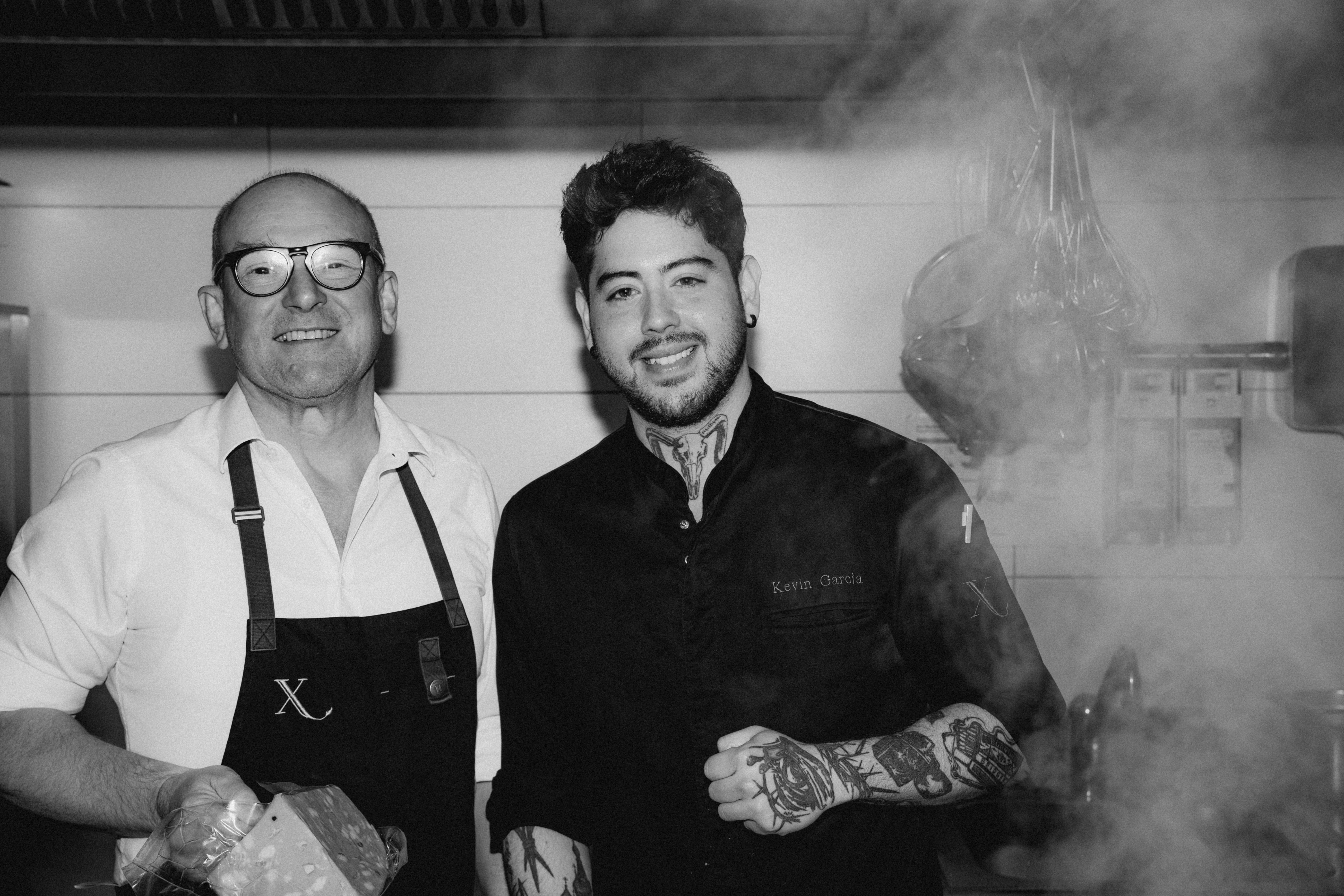 Two persons in a kitchen smiles and gestures while working behind a counter, surrounded by various kitchen equipment.