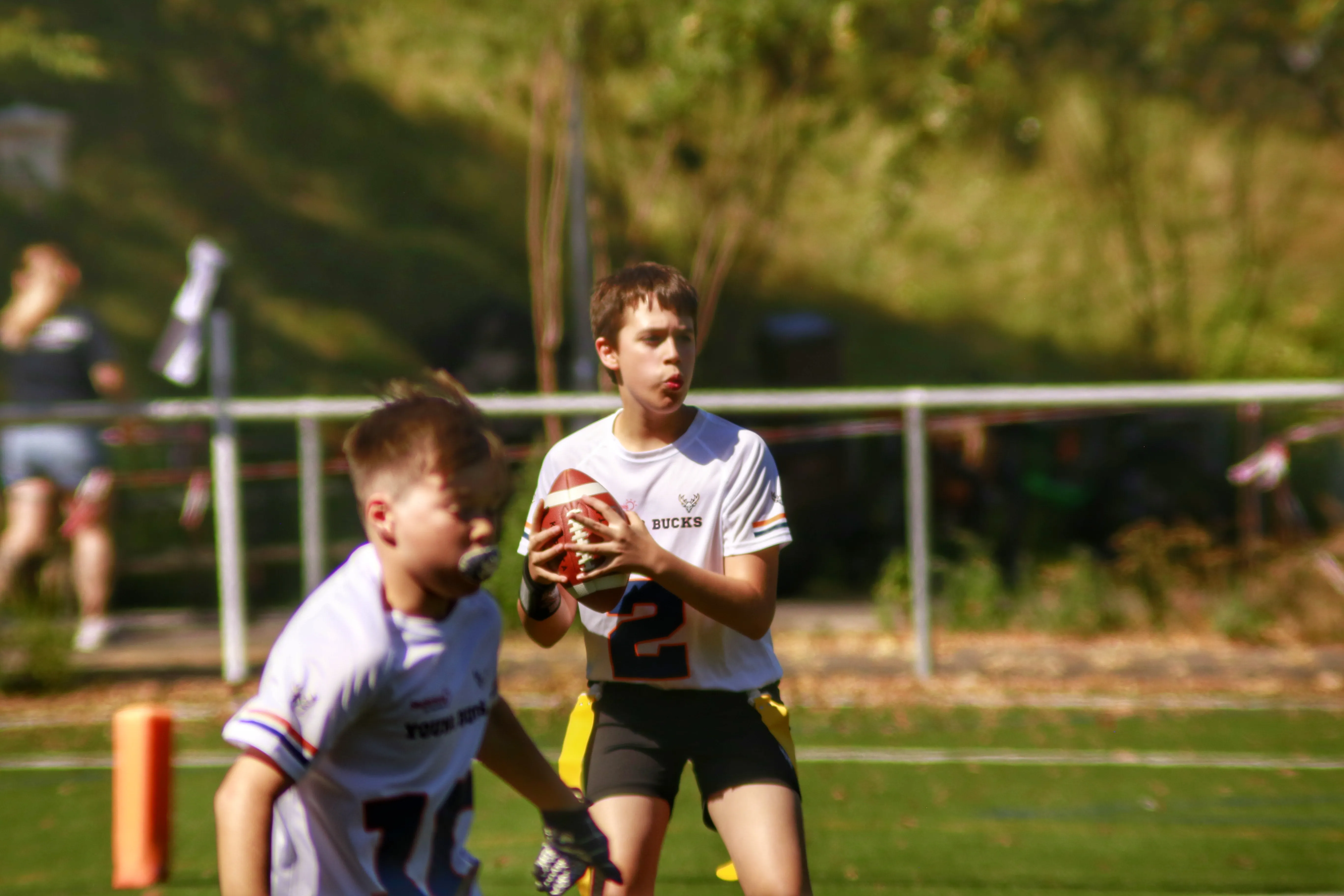 A young athlete prepares to throw a ball while another player looks on, with a grassy field in the background.