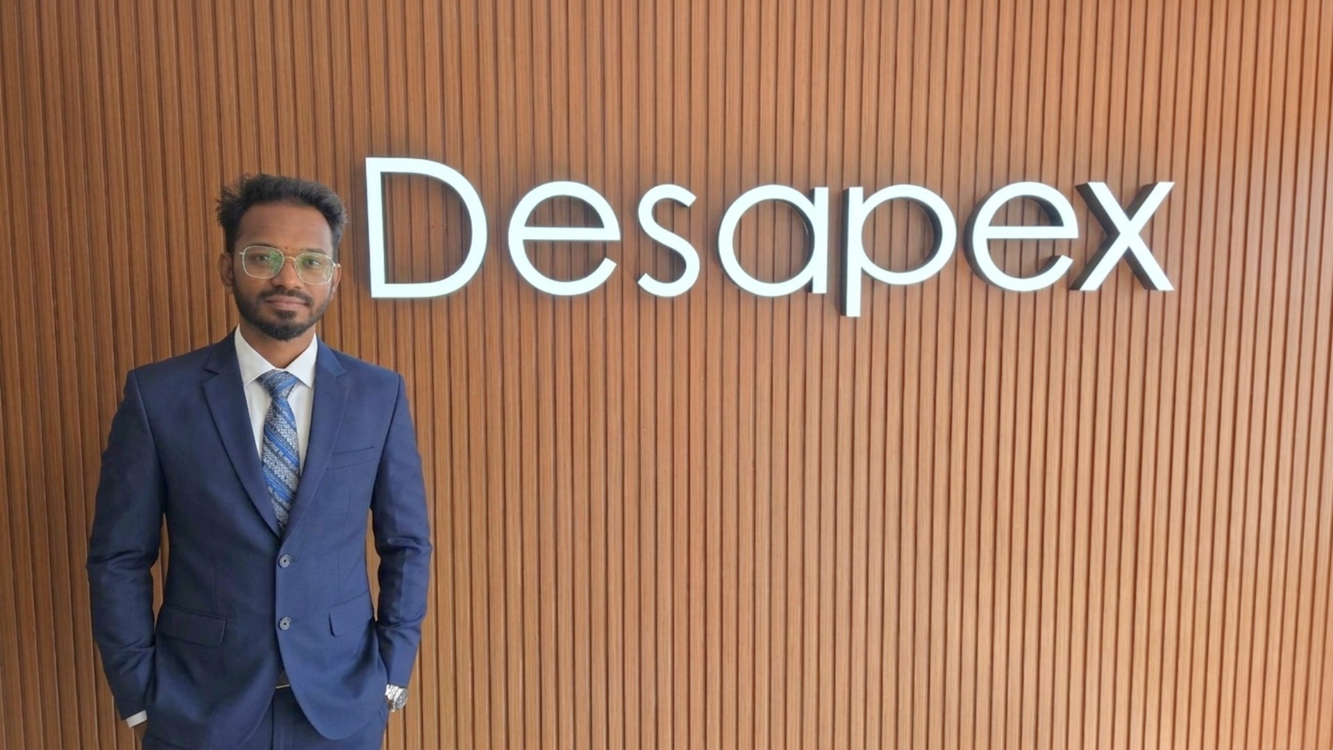 A man stands while two people sit at a table in an office setting, smiling and engaged in conversation. The mood is friendly and collaborative.