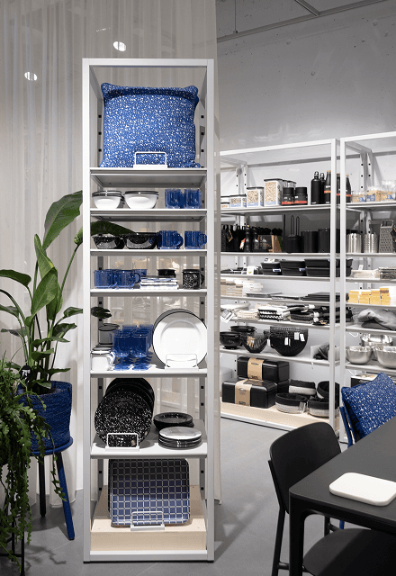 Freistehendes schmales Regalelement des modularen micasa Stores, im Vordergrund ein angeschnittener Tisch und Stuhl, im Hintergrund ein textiler Vorhang.