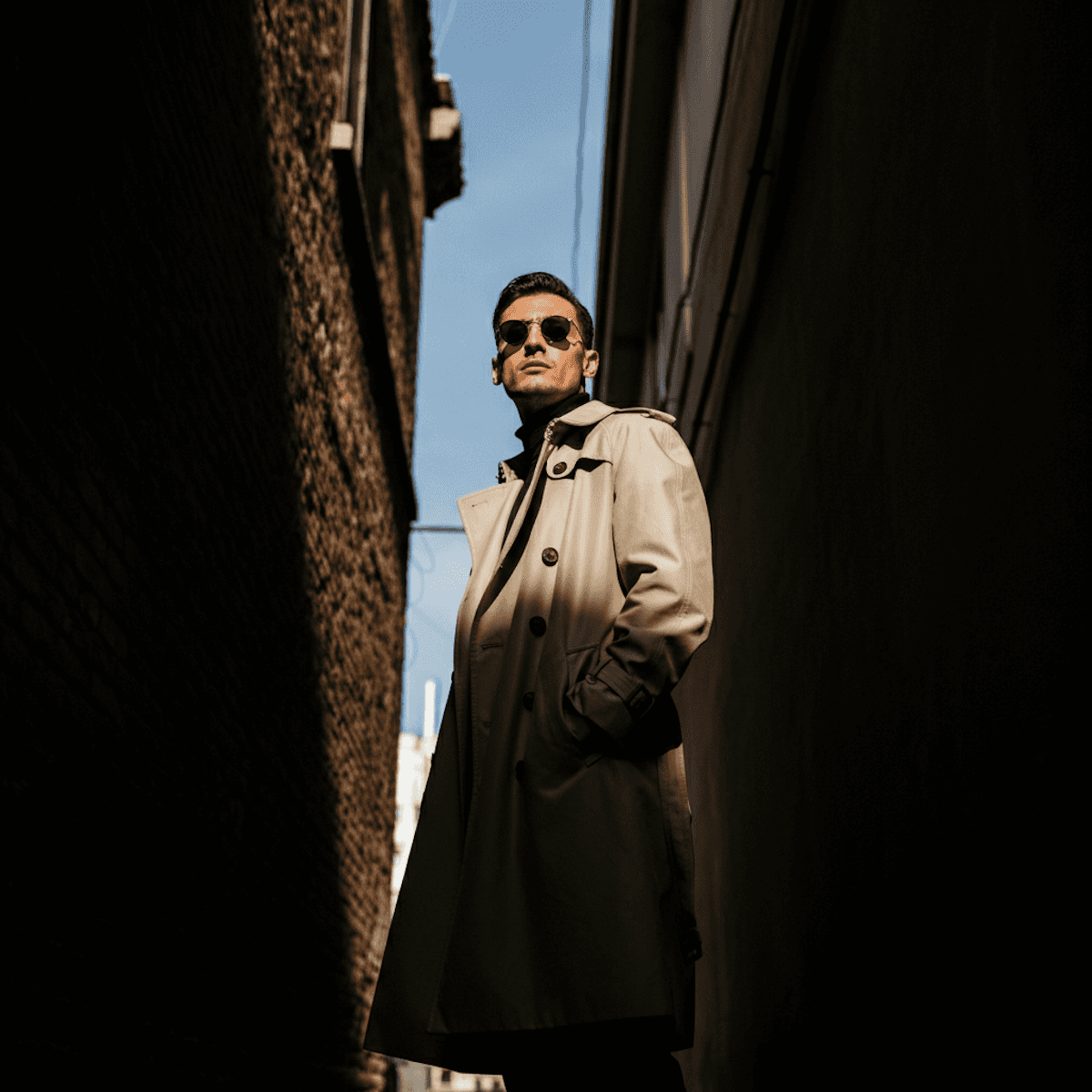 Man in a beige trench coat and sunglasses standing in a narrow alley looking upward.